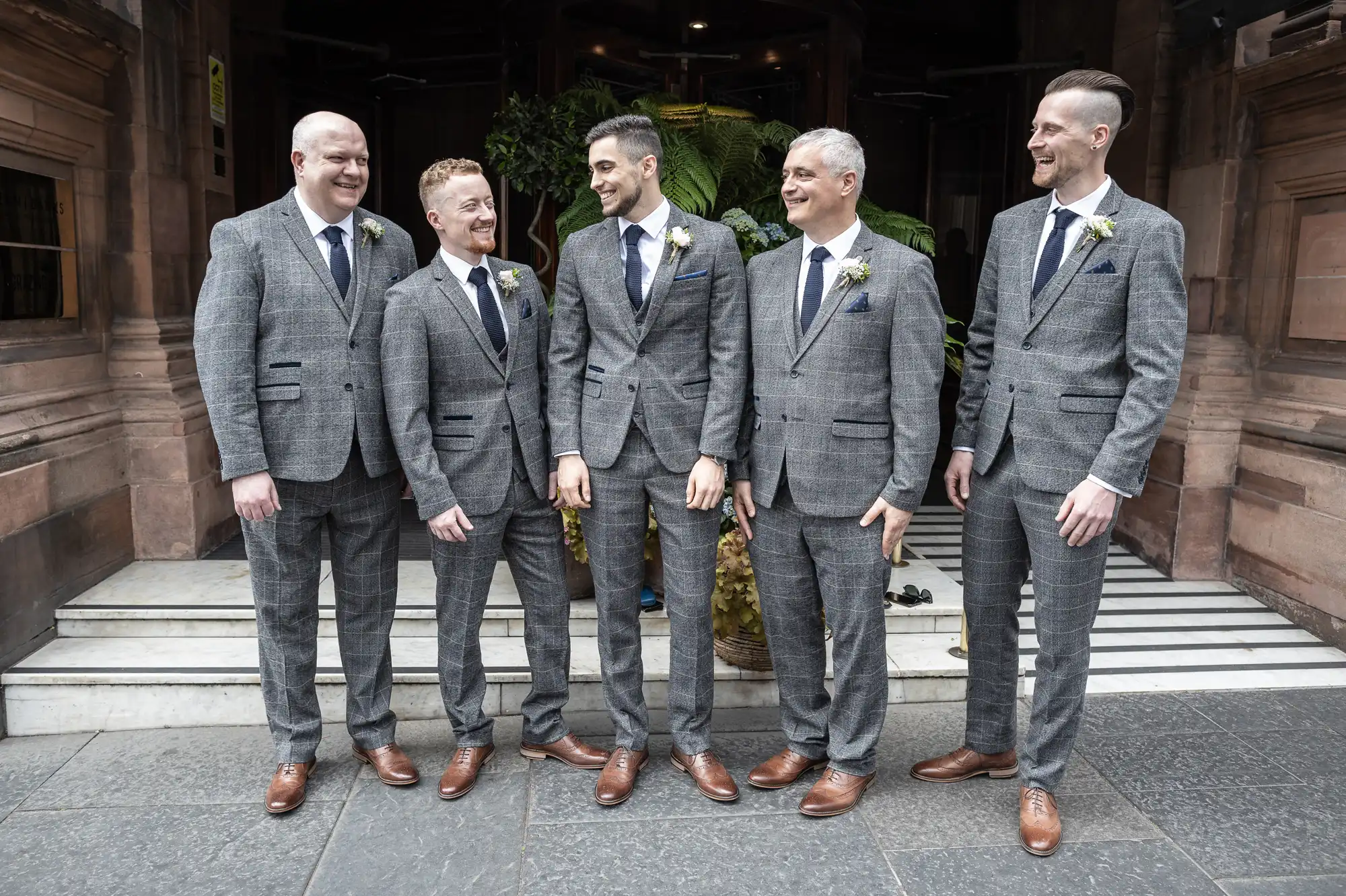 Five men in matching gray suits and brown shoes, standing on steps and smiling, each with a boutonniere on their lapels.
