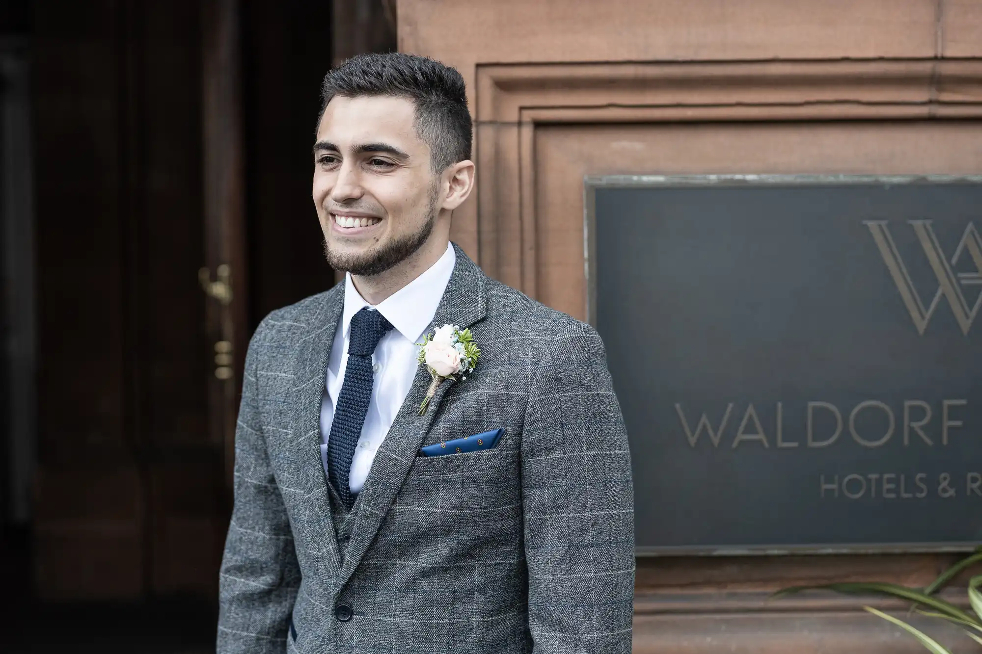 A man in a gray suit and tie, smiling, stands in front of a Waldorf Hotel sign.