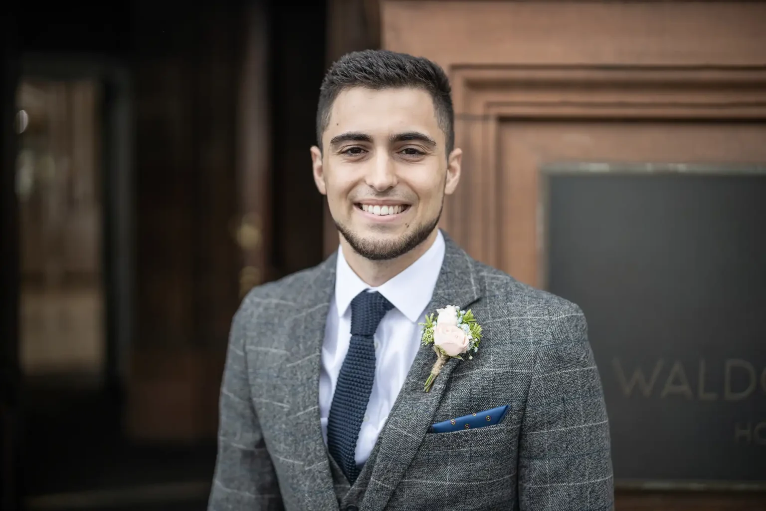 Young man in a gray suit and tie smiling outside a building with a "Waldorf" sign.
