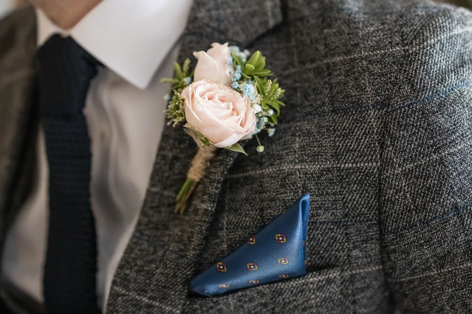 Close-up of a groom's grey tweed suit with a pink rose boutonniere and a blue pocket square with floral patterns.