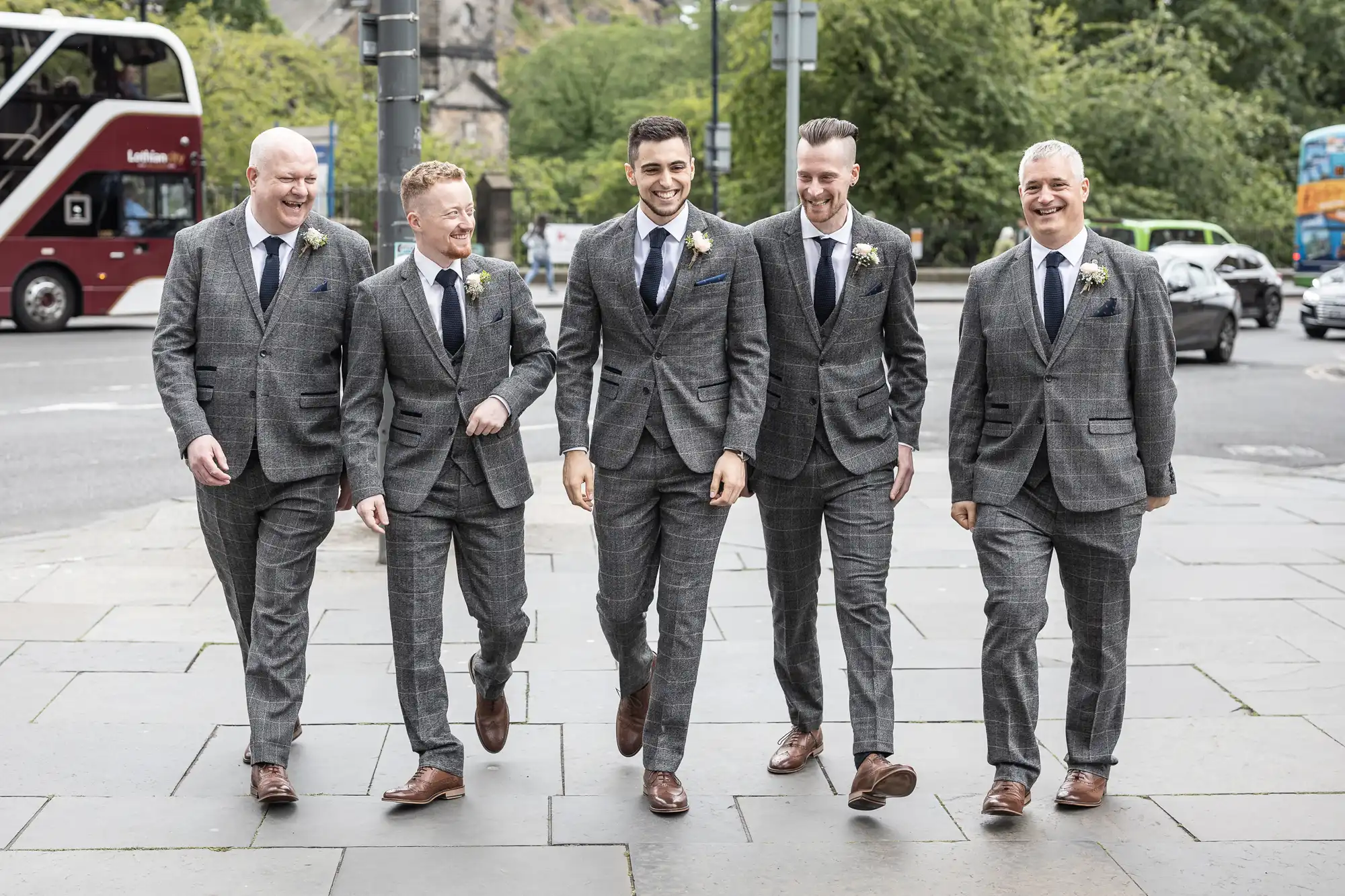 Five men in matching gray suits and boutonnieres walk together smiling on a city sidewalk, with a bus and greenery in the background.
