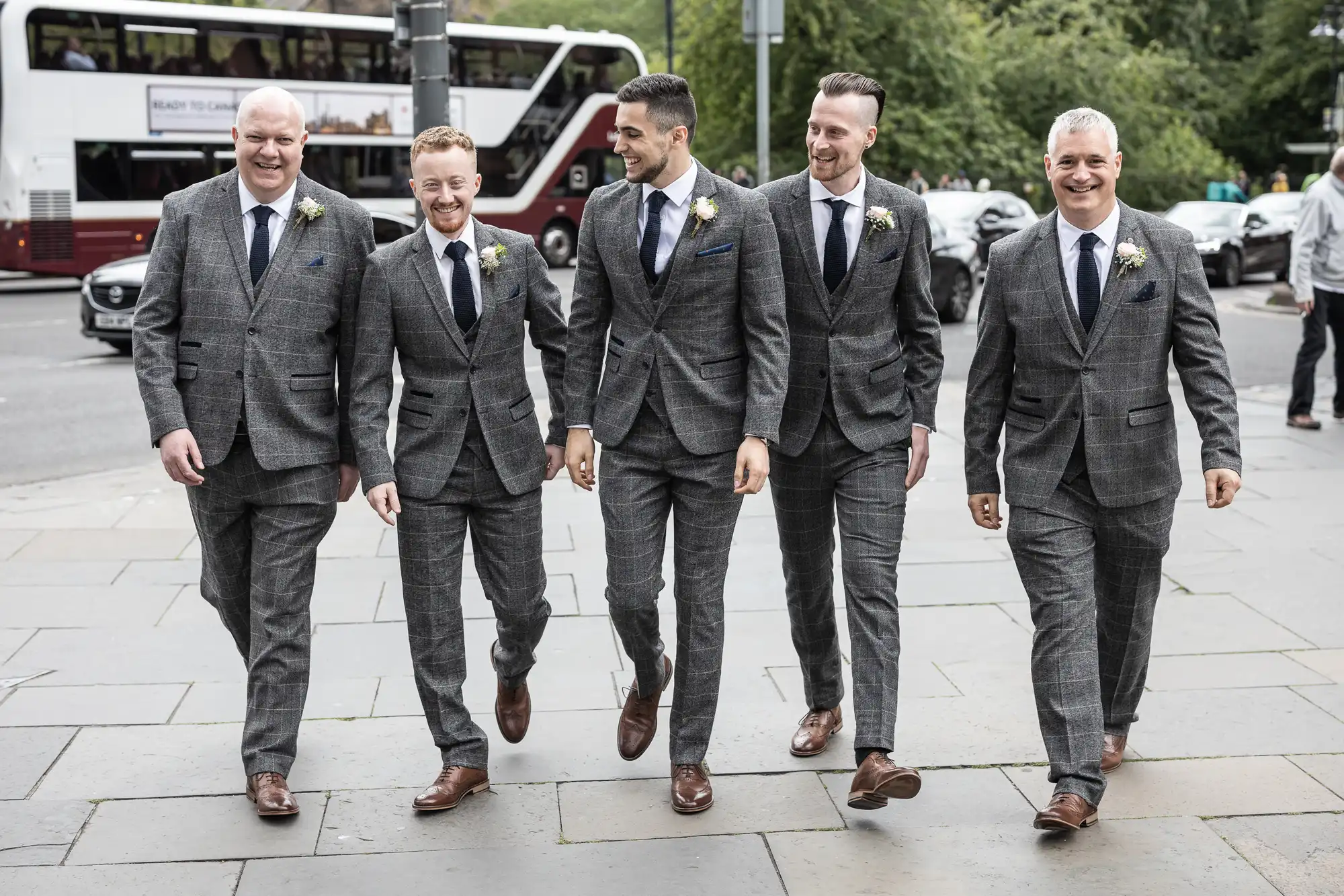 Five men in matching gray suits and boutonnieres walk together and smile on a city sidewalk, with buses in the background.