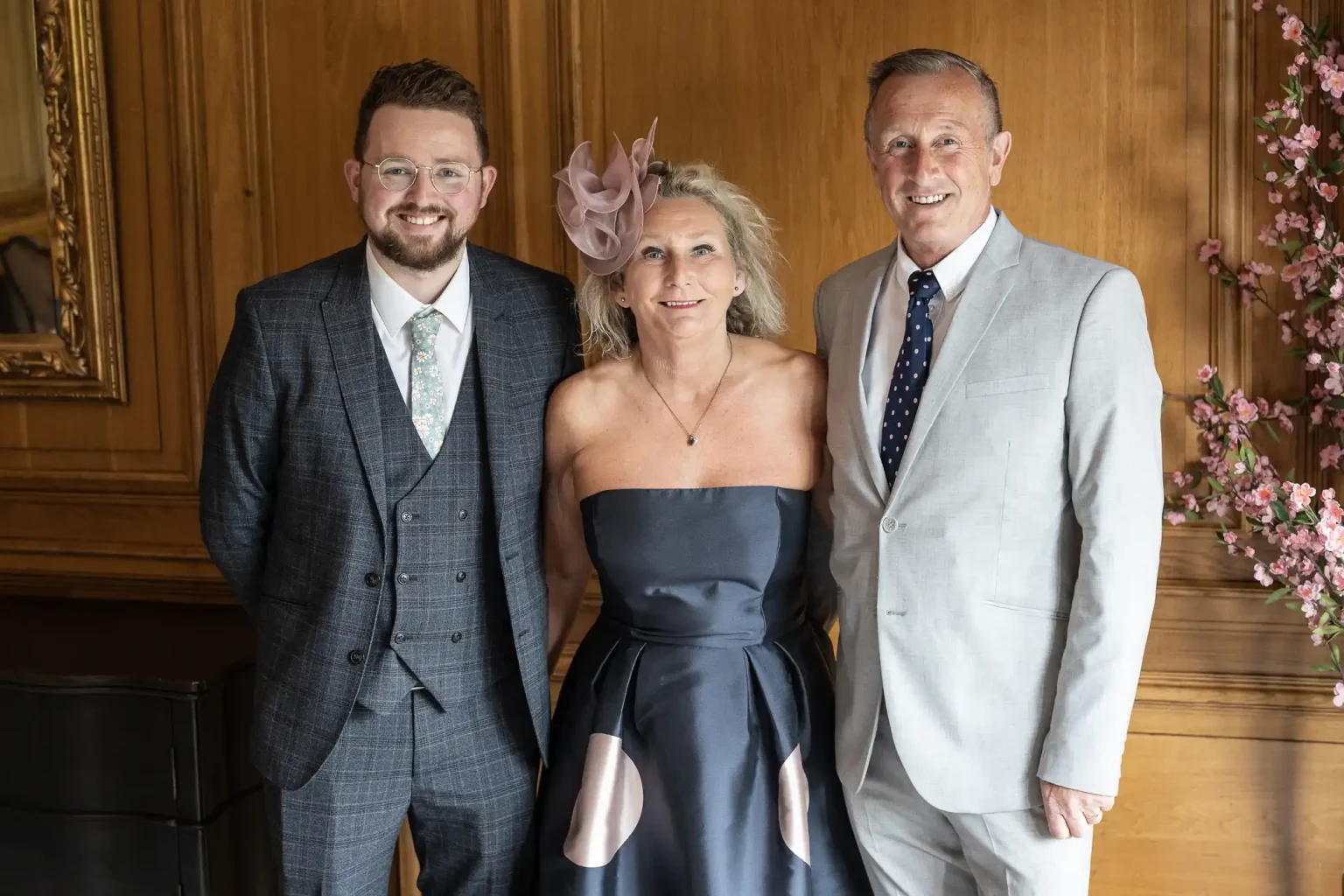 Three adults dressed in formal attire, with two men and one woman, smiling and posing for a photo in an elegant room with wooden paneling and floral decorations.