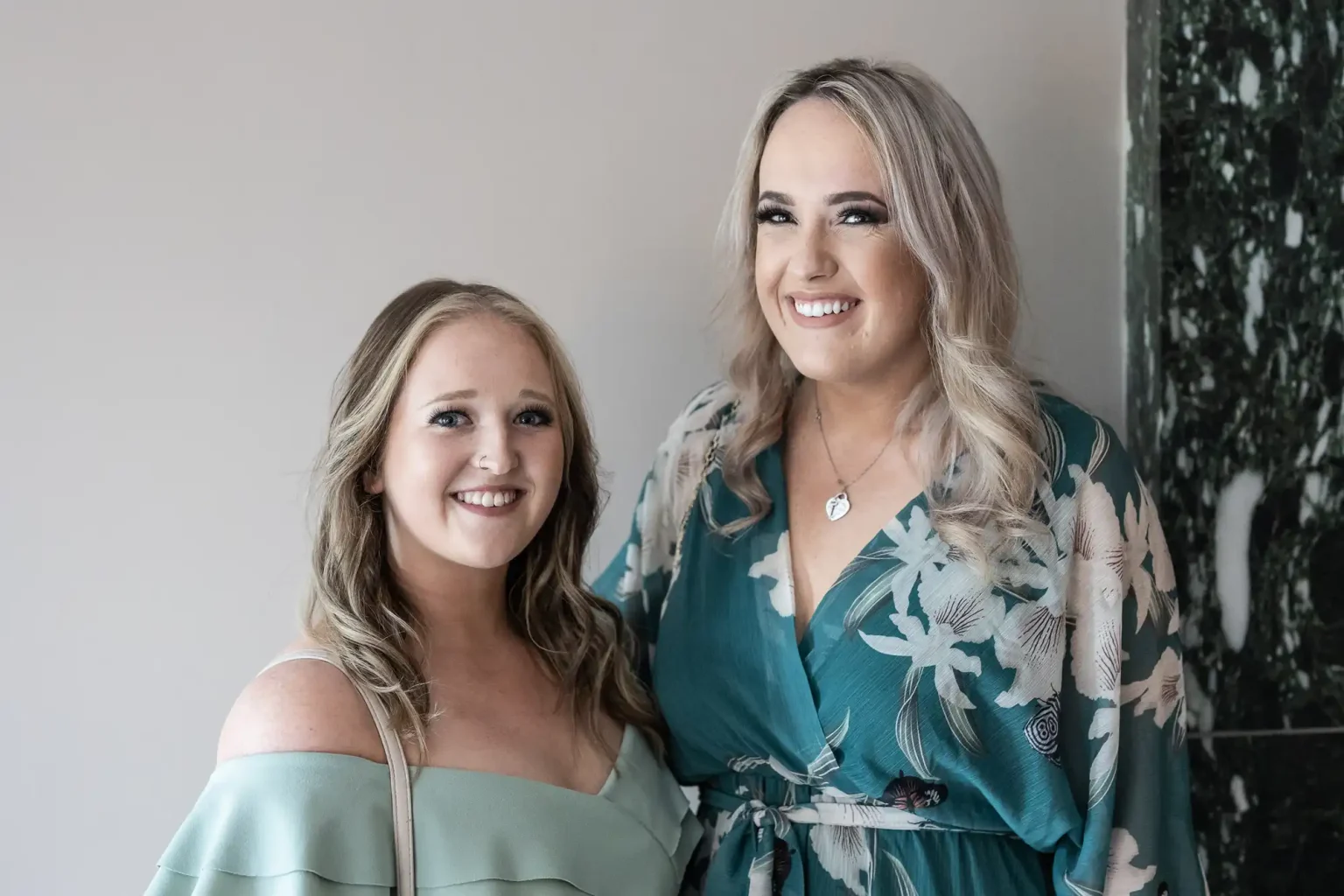 Two women smiling, standing indoors. One wears a green off-shoulder top, and the other wears a floral robe.