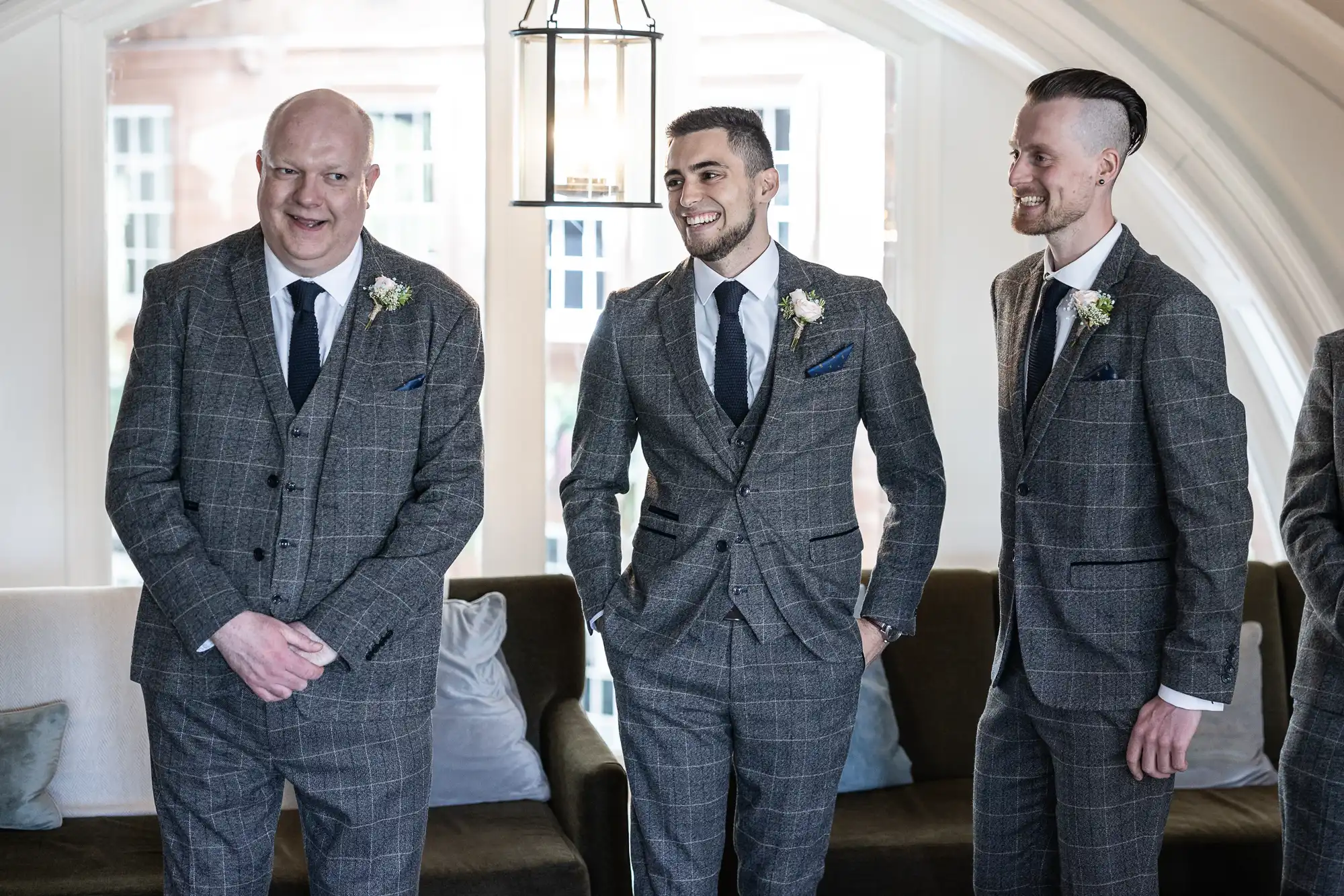 Three smiling men in matching gray suits and boutonnieres standing in a bright, elegant room.
