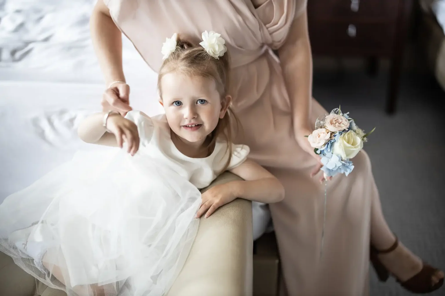 A young girl in a white dress sits on a woman's lap, holding a small bouquet, both smiling.