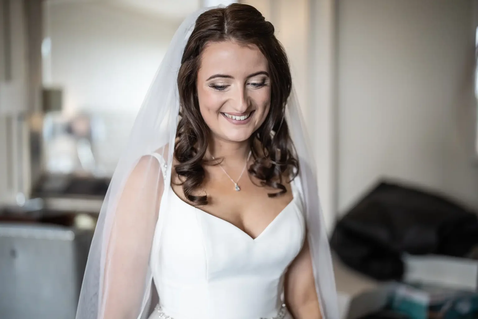 A bride in a white dress and veil smiles gently, looking down in a softly lit room.