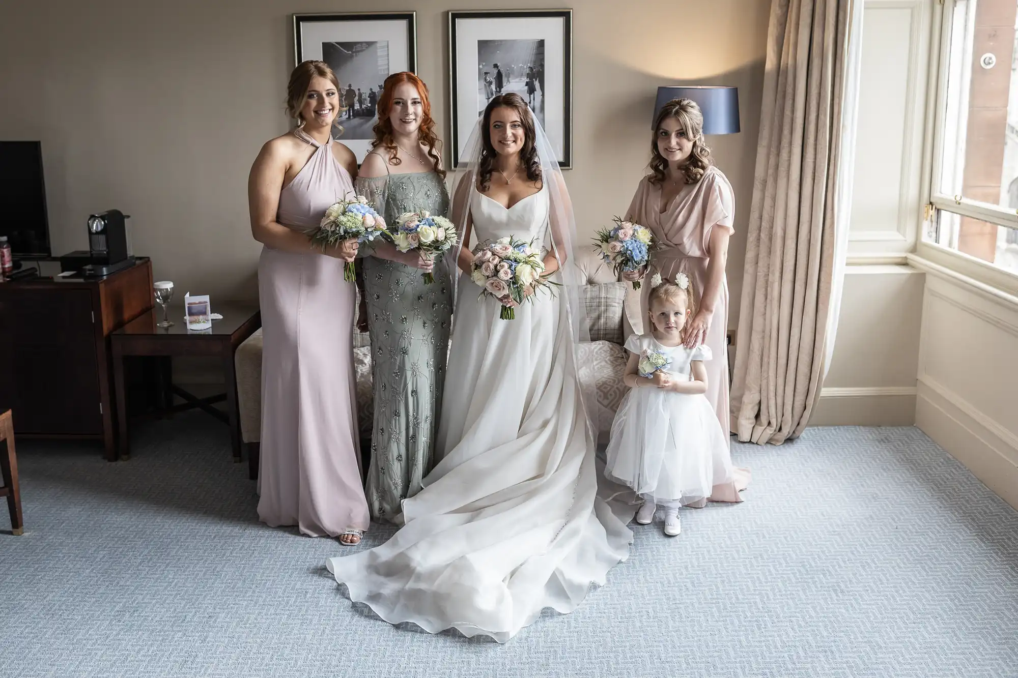 Bride in a white gown with three bridesmaids and a young flower girl, all holding bouquets, posing in a hotel room.