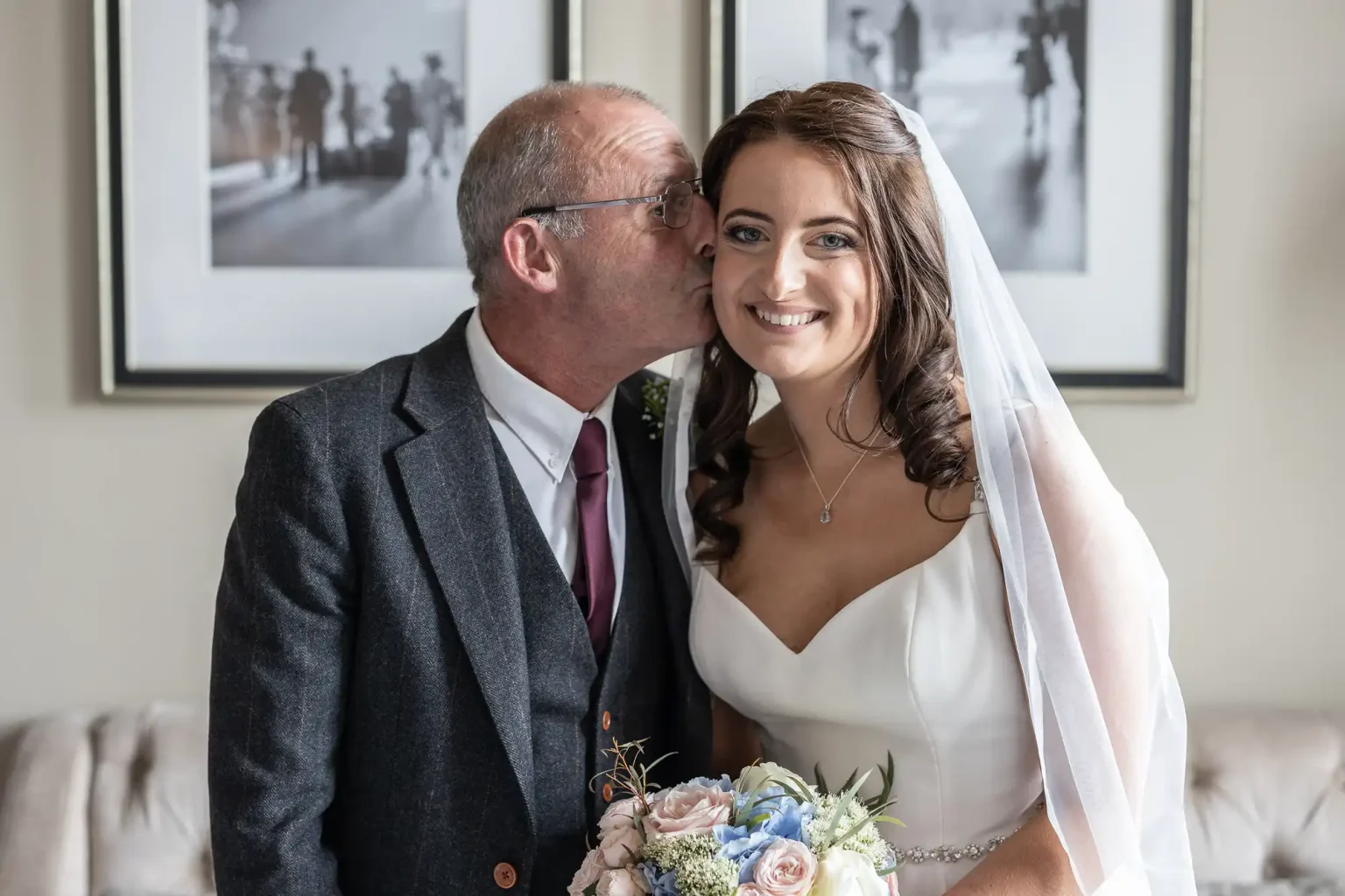 A bride in a white dress and veil, holding a bouquet, smiles next to an older man in a suit as he kisses her on the cheek. Black and white photos adorn the wall behind them.