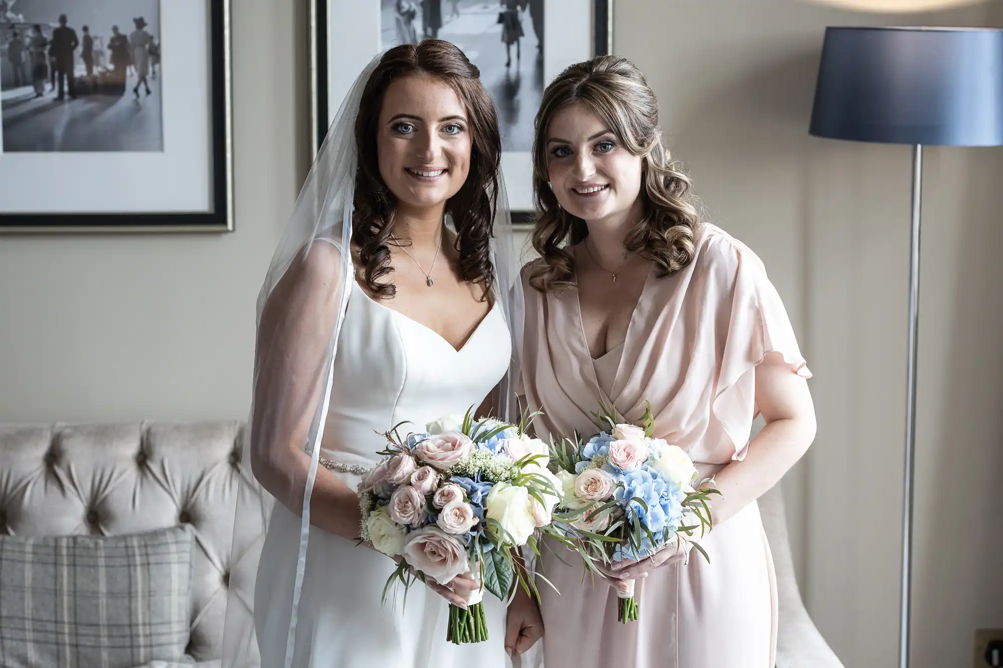 Two women in formal attire, one in a white dress and the other in a pink shawl, holding bouquets and smiling, standing in a room with elegant decor.