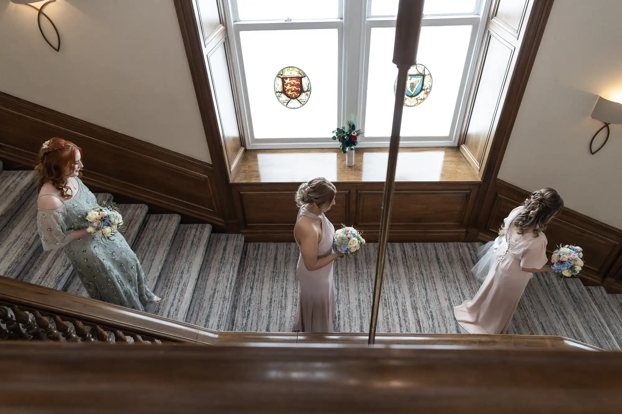 Three bridesmaids in elegant dresses holding bouquets stand on a grand staircase with stained glass windows above.
