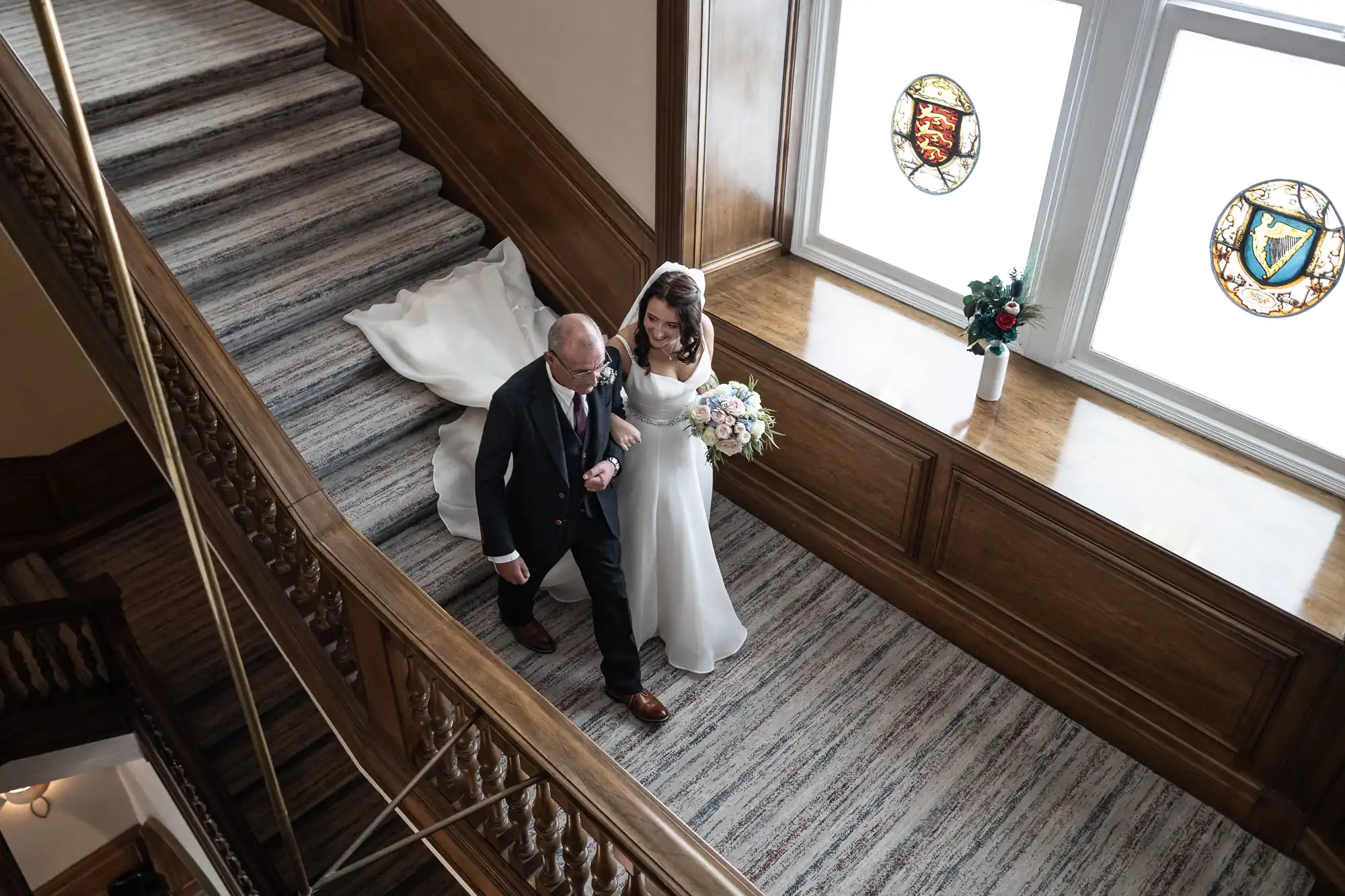 A bride and her father descend a grand staircase, beside stained glass windows, in a classic wedding scene.