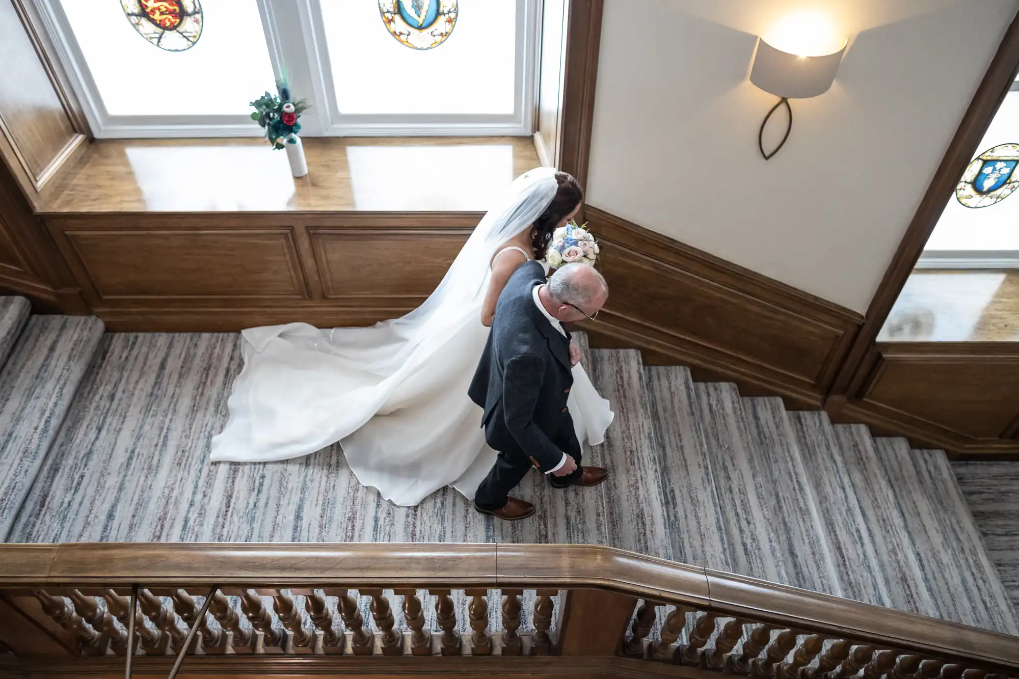 A bride with a flowing white dress walks arm in arm with an elderly man down a wooden staircase in an elegant building, viewed from above.
