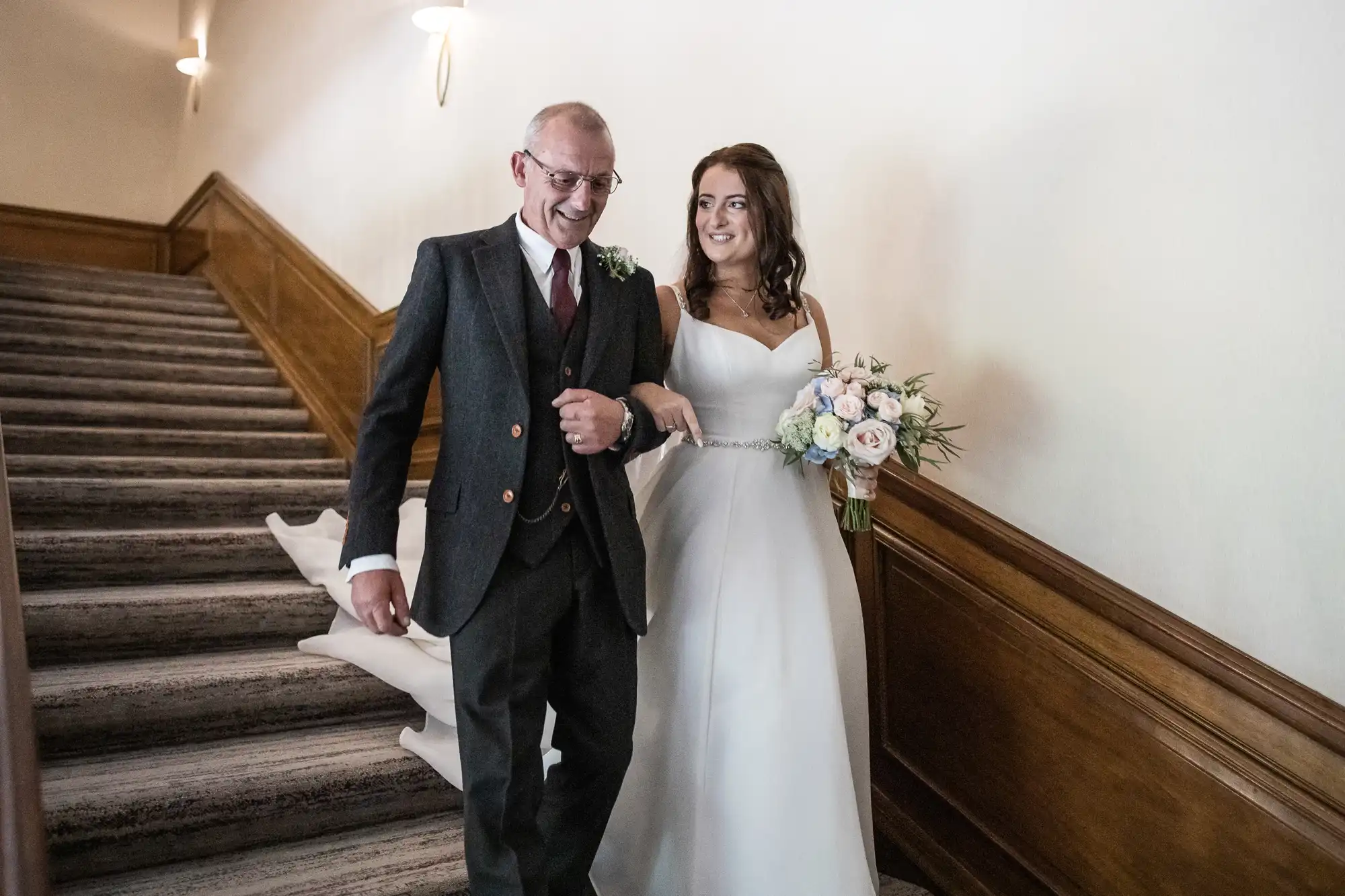 A bride in a white gown and her father in a gray suit and glasses descend a staircase, both smiling, with the bride holding a bouquet of pastel flowers.