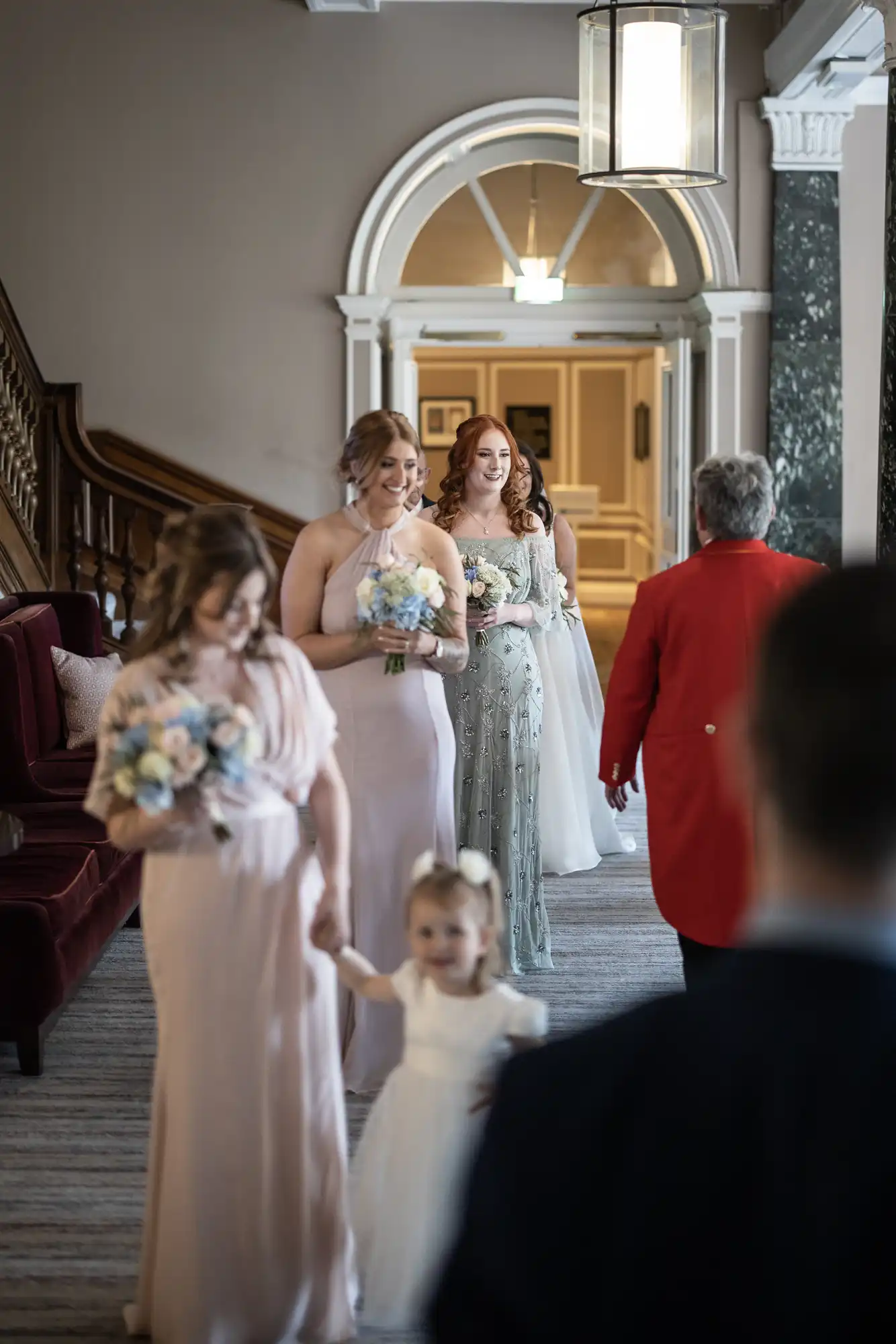 A wedding procession in an elegant hallway with a toddler leading, followed by women in formal dresses holding bouquets.