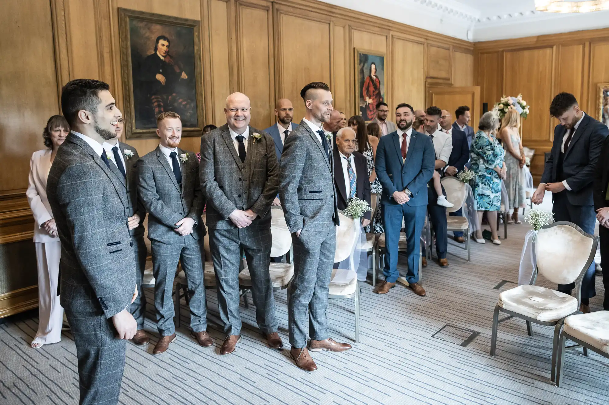 A wedding ceremony in a decorated room with guests and groomsmen standing as a groom awaits at the front.