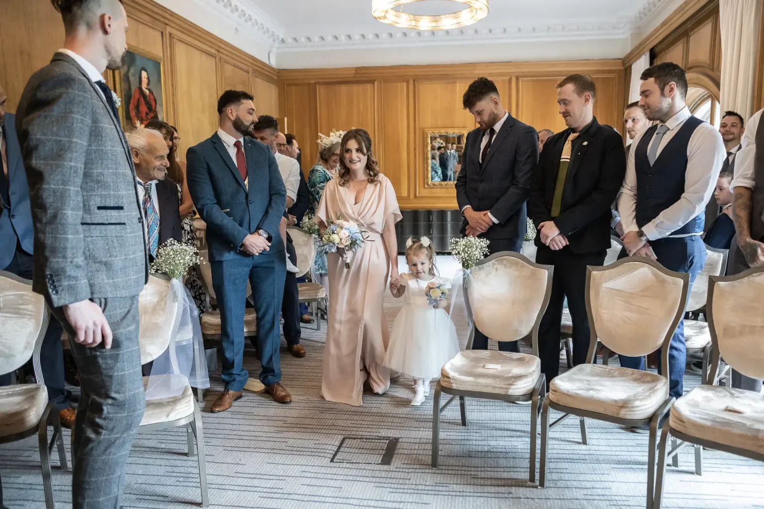 A wedding ceremony in an elegant room, with a bridesmaid walking down the aisle accompanied by two young flower girls, as guests watch attentively.