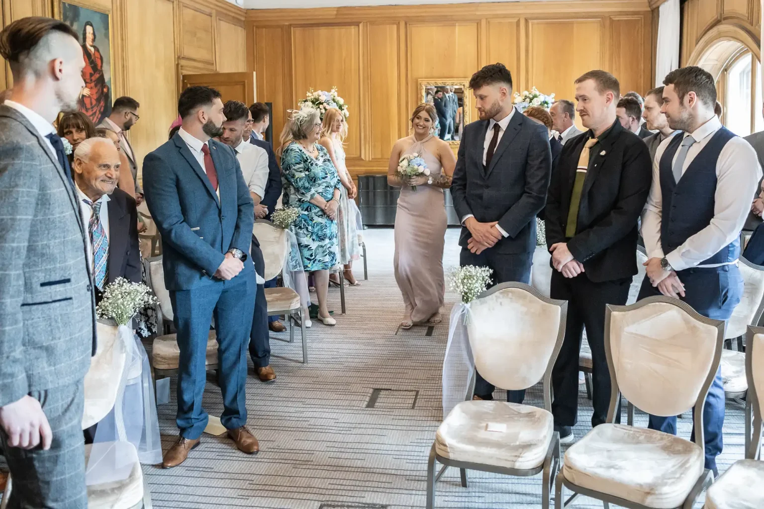 A bridesmaid walking down the aisle, holding a bouquet, while guests and groomsmen, standing to one side, watch her intently in a decorated room.