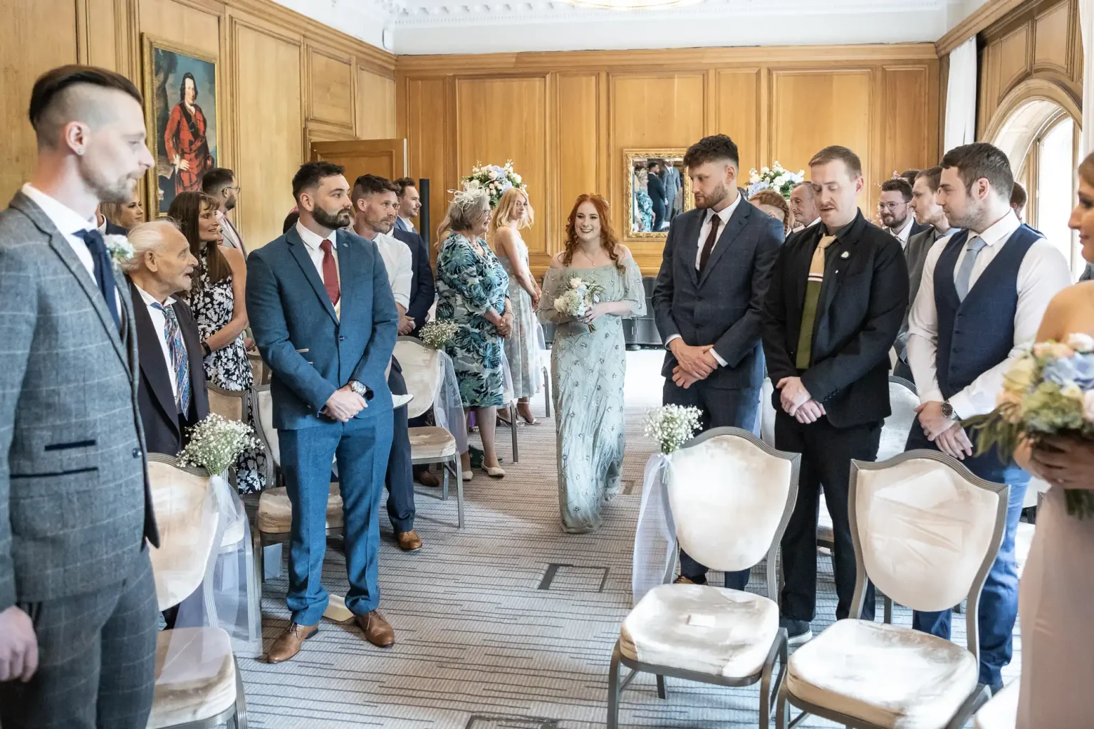 A bridesmaid walks down the aisle in a room filled with guests, accompanied by a man, both smiling during a Western-style wedding ceremony.