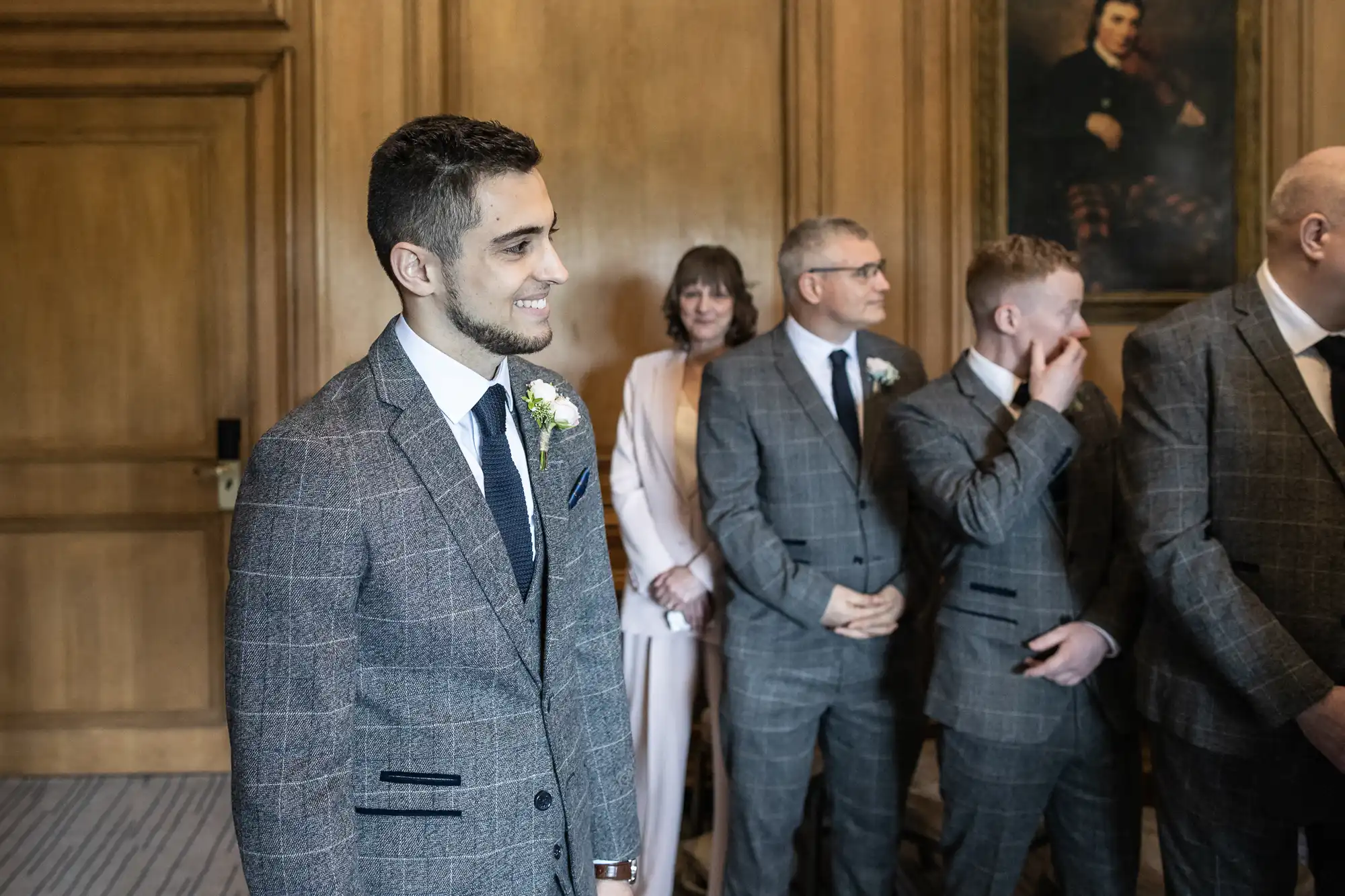 A groom in a gray plaid suit smiles during a wedding ceremony in an elegant room, with four guests in the background.