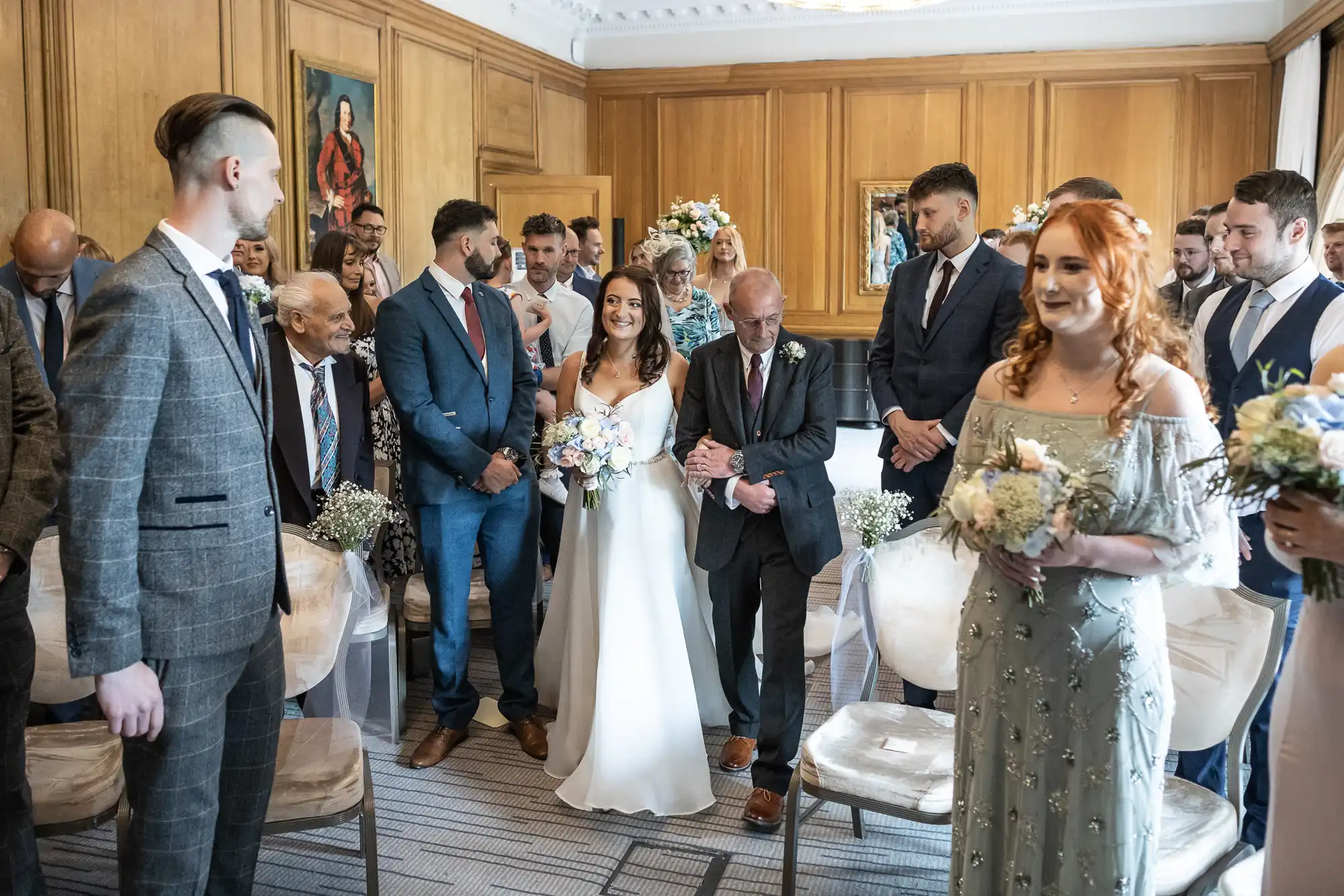 A wedding ceremony in a decorated room with guests watching as the bride walks down the aisle accompanied by an older man.