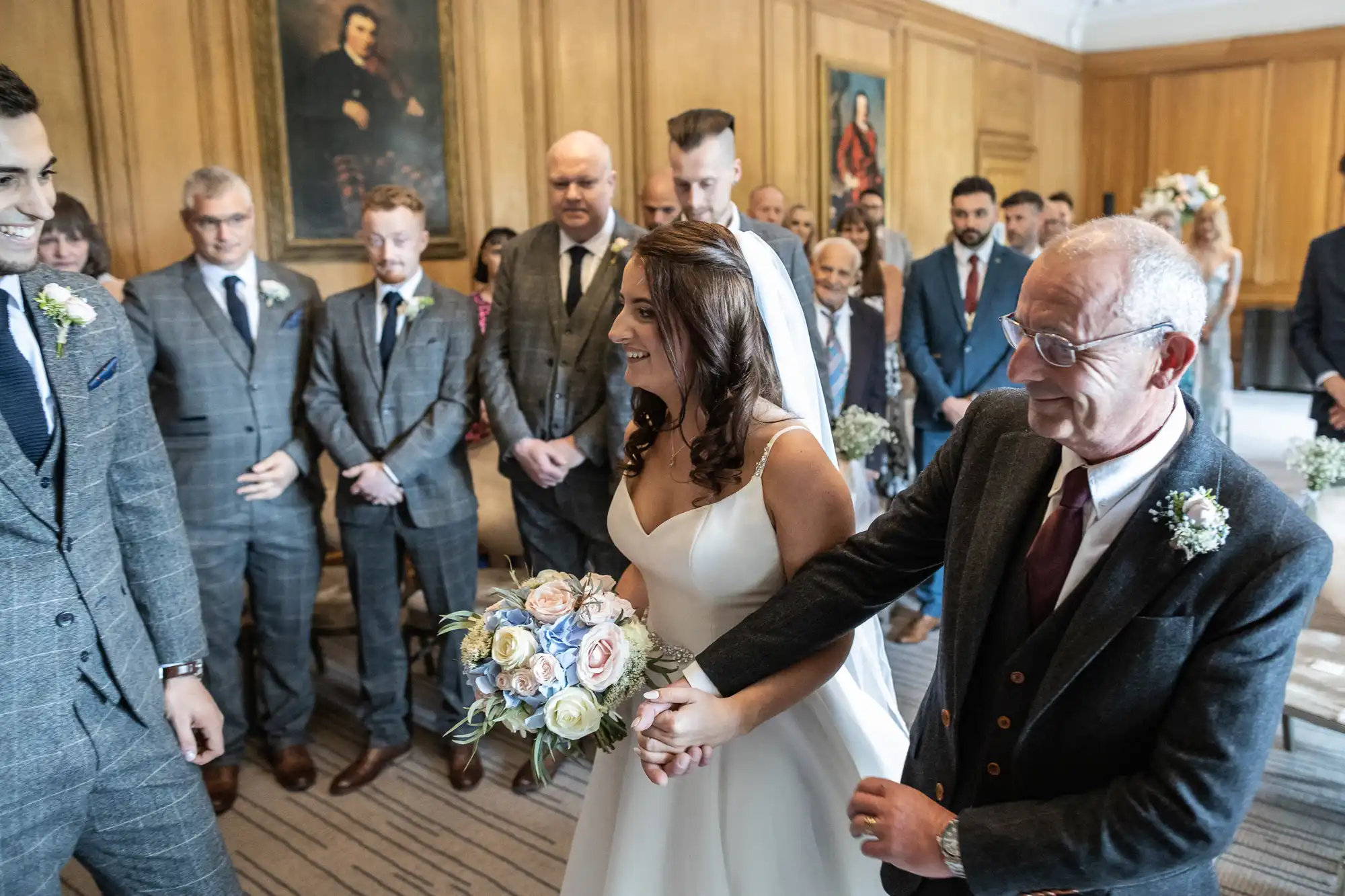 A bride and her father walking down the aisle, with guests looking on, in an indoor ceremonial setting.
