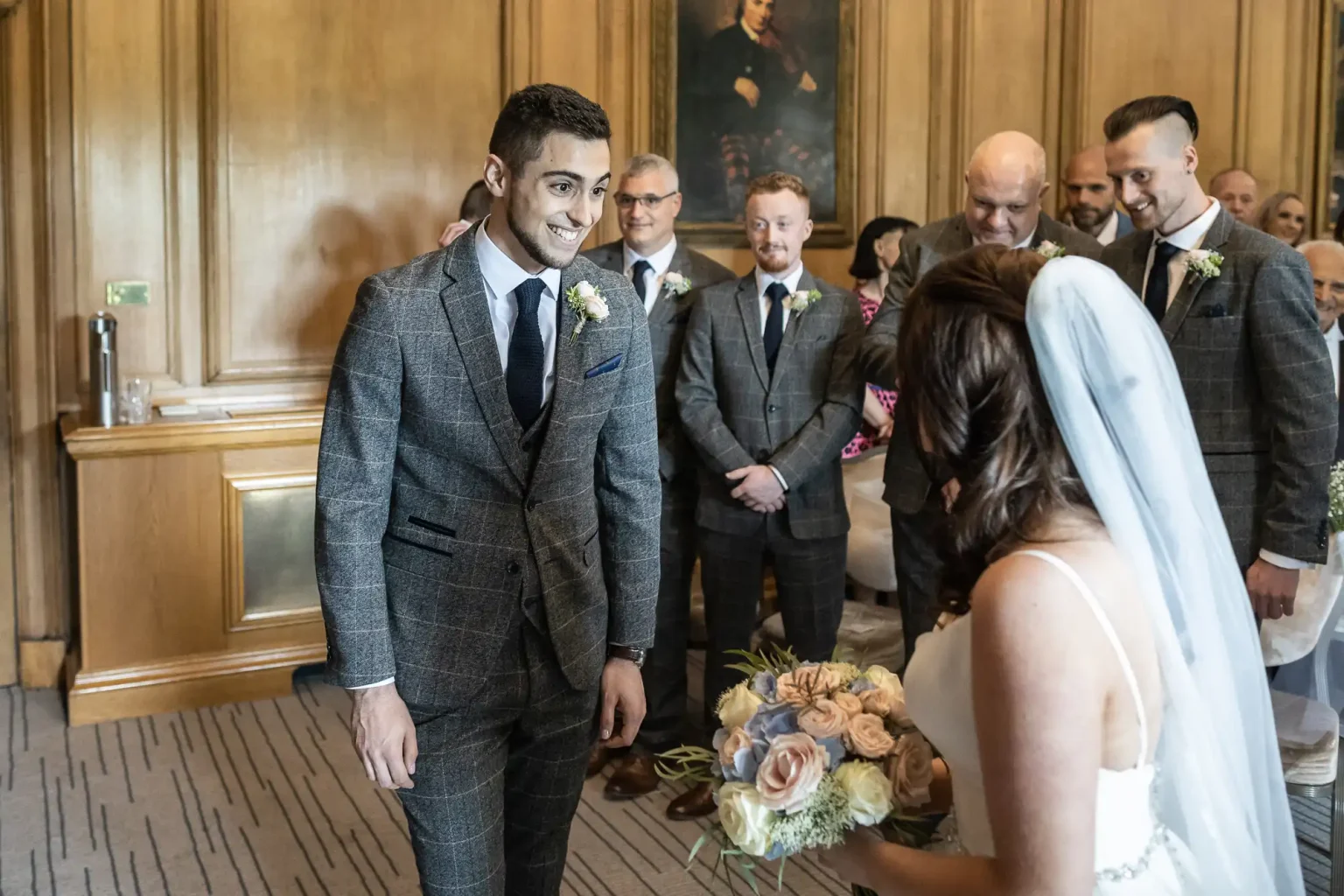 Groom in a gray suit smiling at bride holding a bouquet, surrounded by wedding guests in a wood-paneled room.