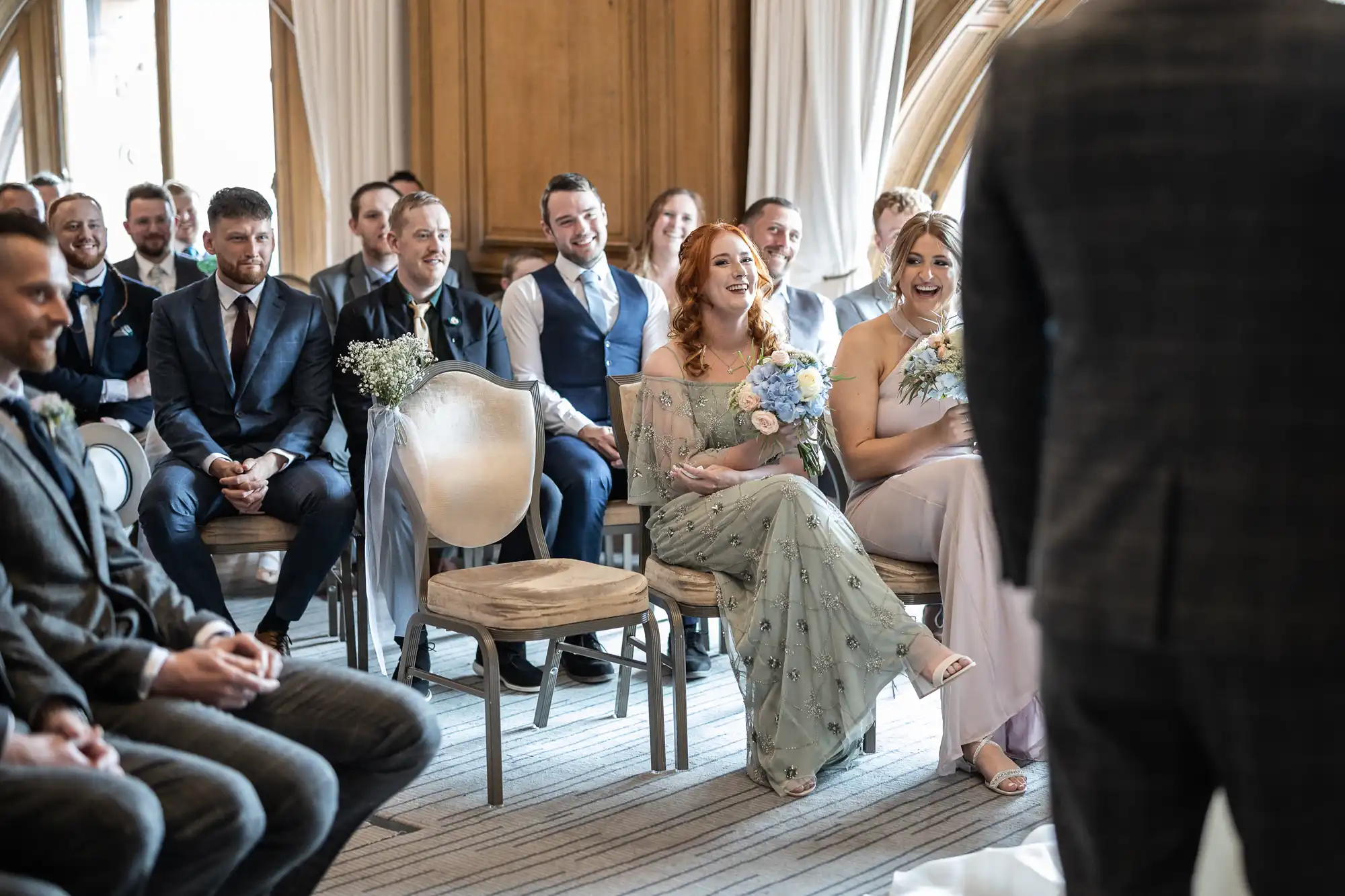 A wedding ceremony in a bright room with guests smiling at a bride, who is seated in the center holding a bouquet.