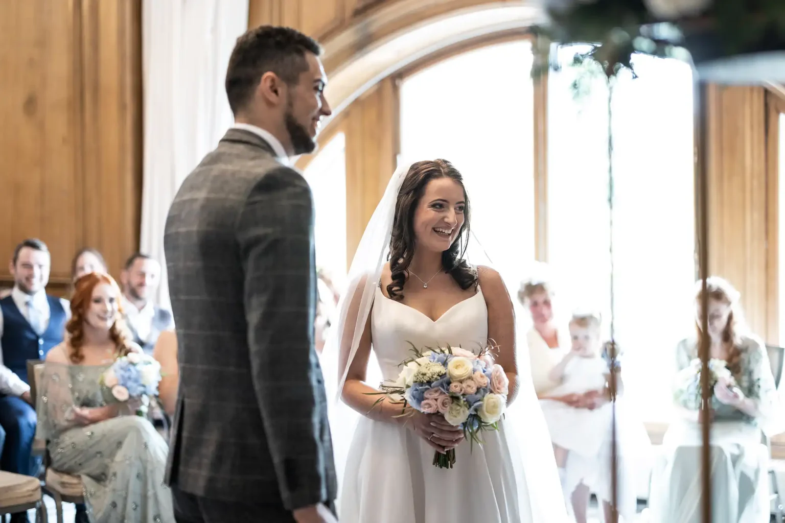 Bride smiling at groom in a sunlit room, holding a bouquet, with guests seated in the background.