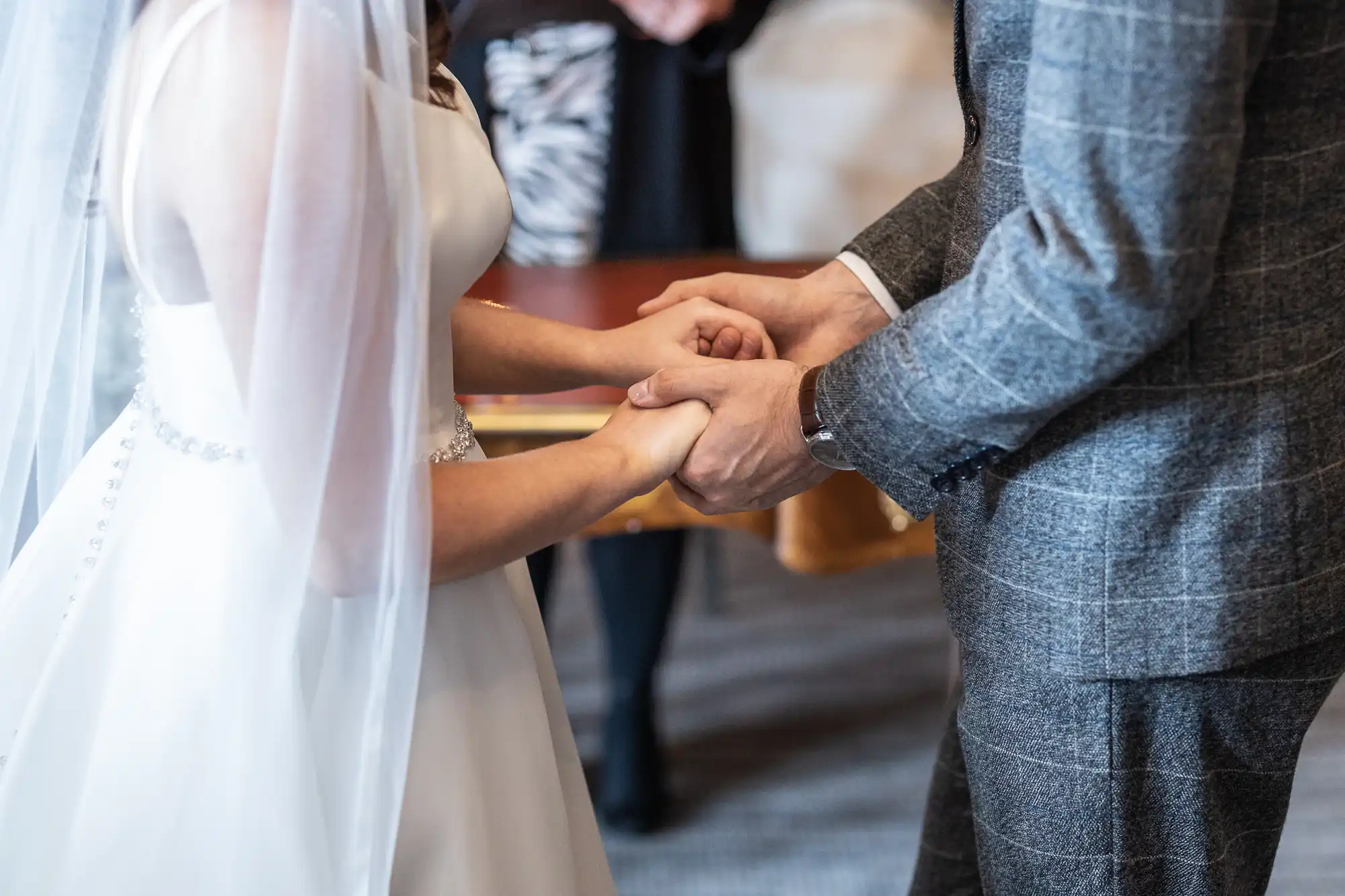 A bride in a white dress and a groom in a gray suit hold hands during a wedding ceremony.
