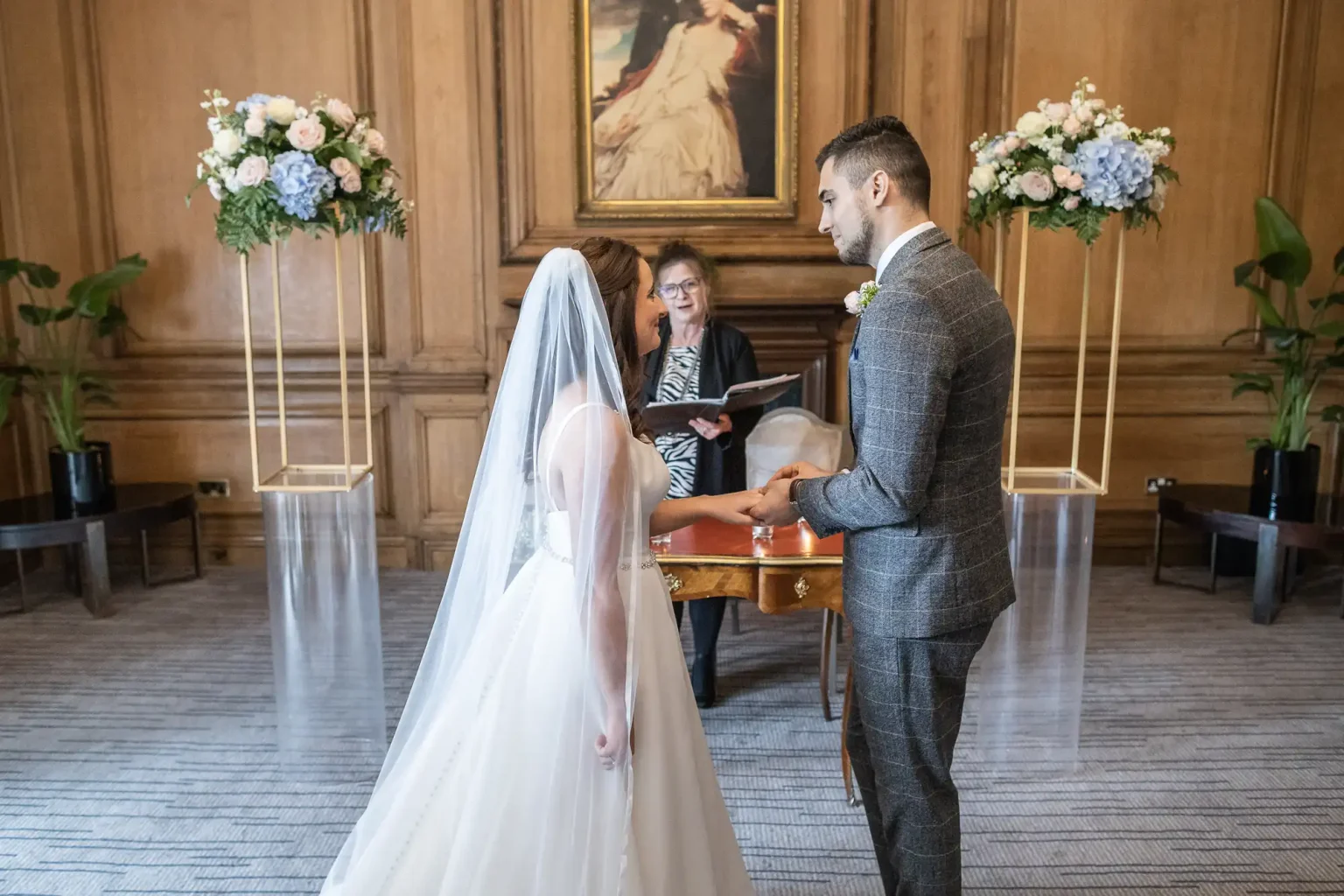 A bride and groom exchange vows in front of an officiant at a grand indoor ceremony, flanked by floral arrangements.