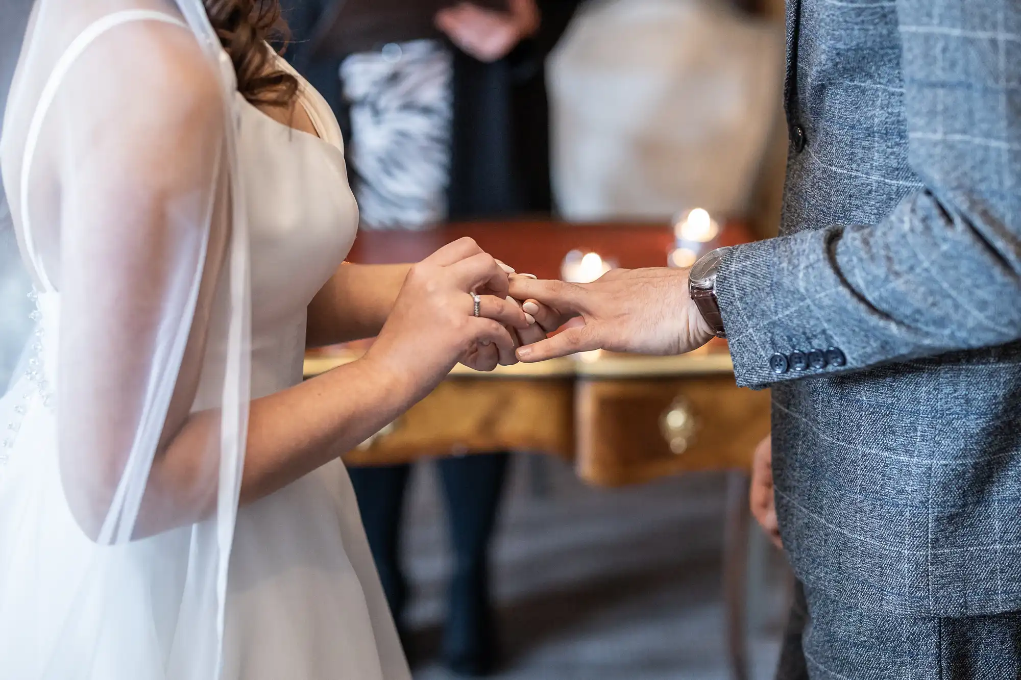 Bride placing a wedding ring on groom's finger during ceremony, close-up of hands with candles in background.