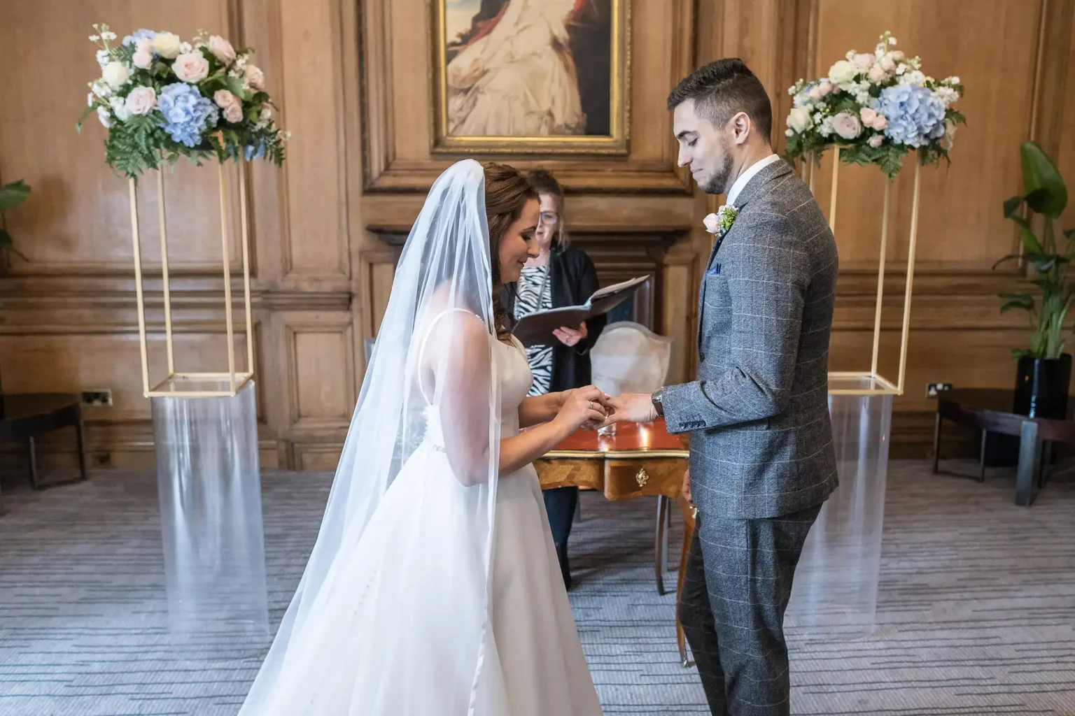 A bride and groom exchange rings at a wedding ceremony, officiated by a woman, in an elegant room with floral decorations.