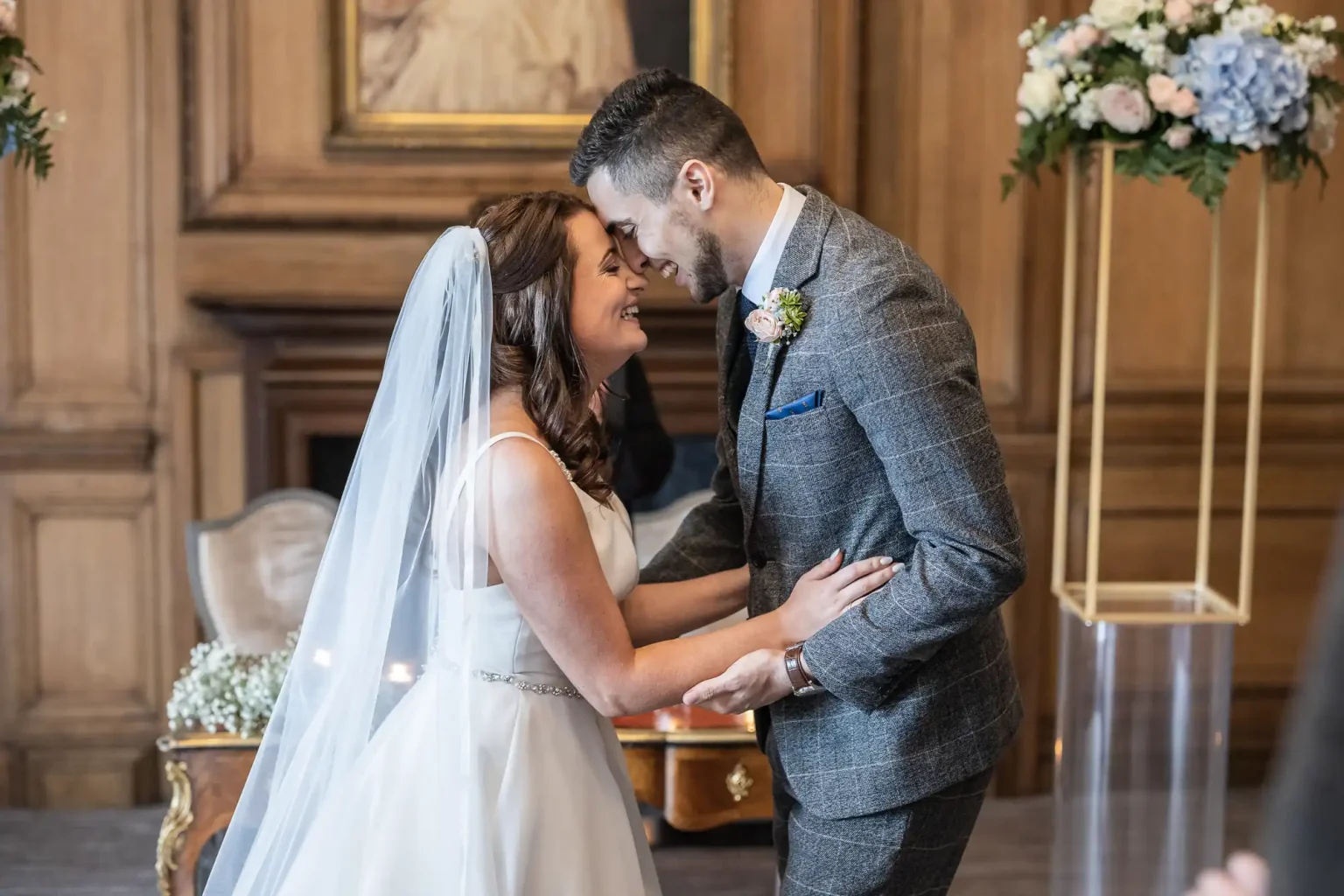 A bride and groom holding hands and touching foreheads, smiling during their wedding ceremony in an elegant room.
