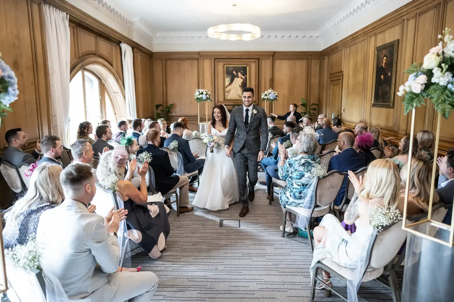 A bride and groom walking down the aisle, smiling at their guests in a well-decorated room with elegant wood paneling.