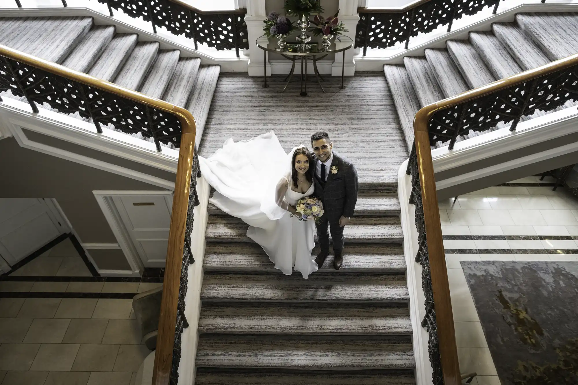 A bride in a long white dress and a groom in a suit descend a grand staircase, smiling, in an elegant lobby.