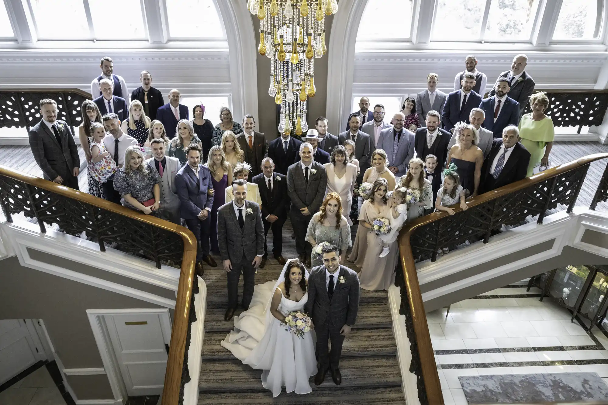 A bride in a long white dress and a groom in a suit on the grand staircase, smiling, in an elegant lobby surrounded by family and friends during the group photo of everyone