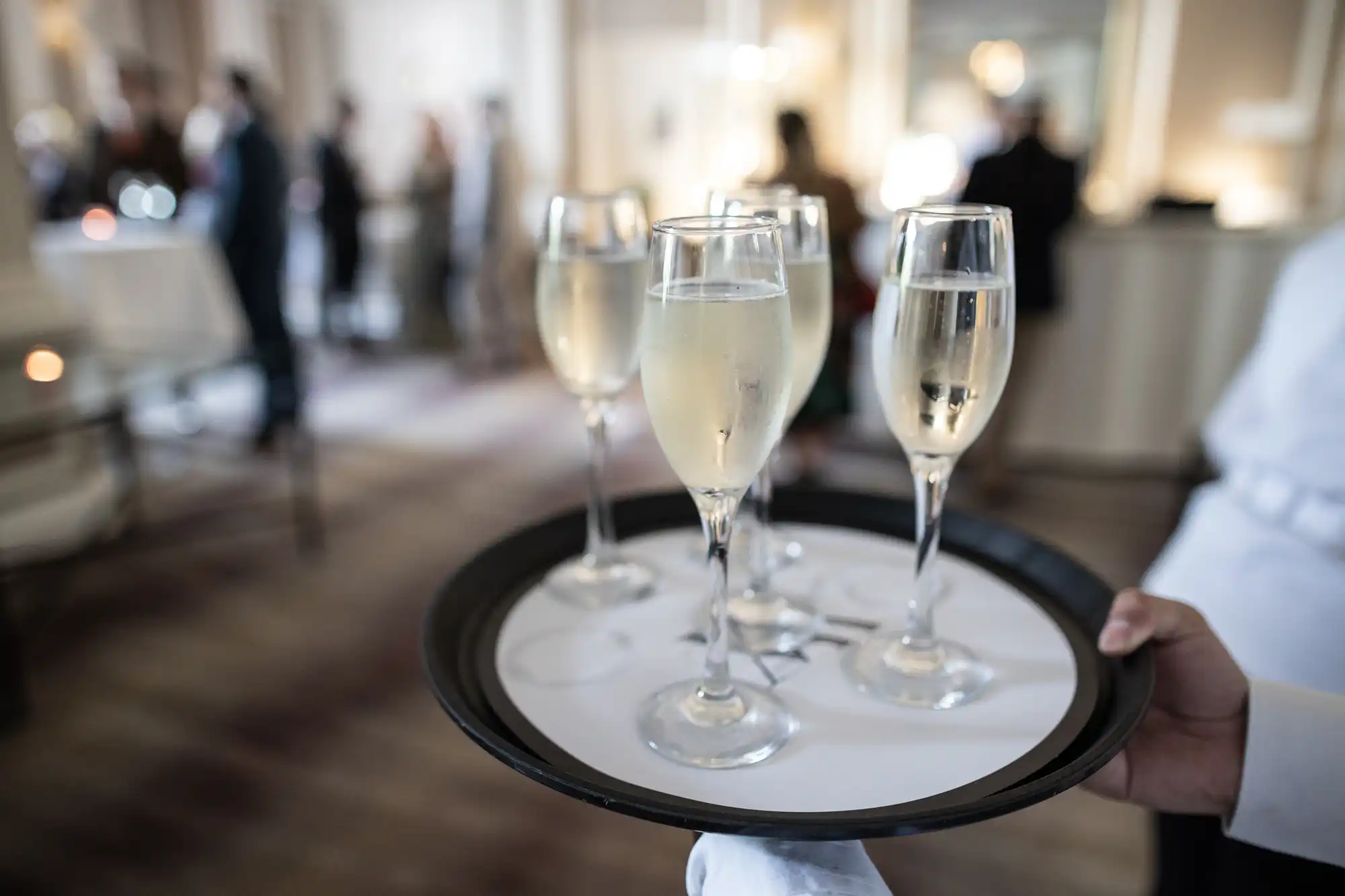 Server holding a tray of champagne glasses at a social gathering in an elegant room with guests in the background.