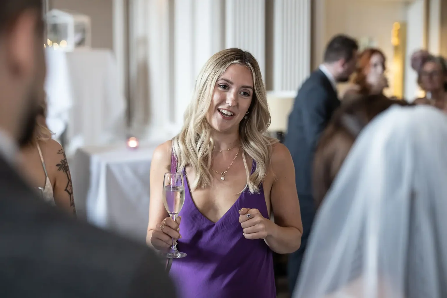 A woman in a purple dress, smiling and holding a glass of champagne at a social event, with other guests in the background.