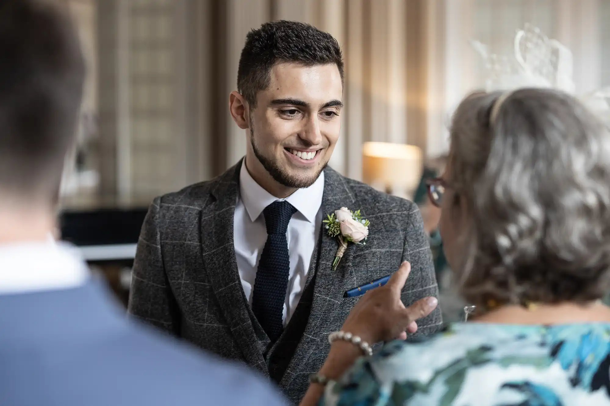A smiling groom in a gray suit and boutonniere converses with wedding guests in an elegant room.