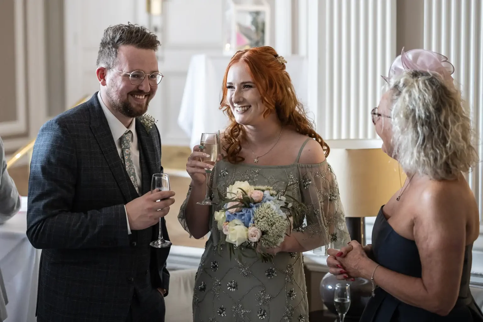 A man and two women chatting indoors at a social gathering, one holding a glass and another holding a floral bouquet, all are smiling.