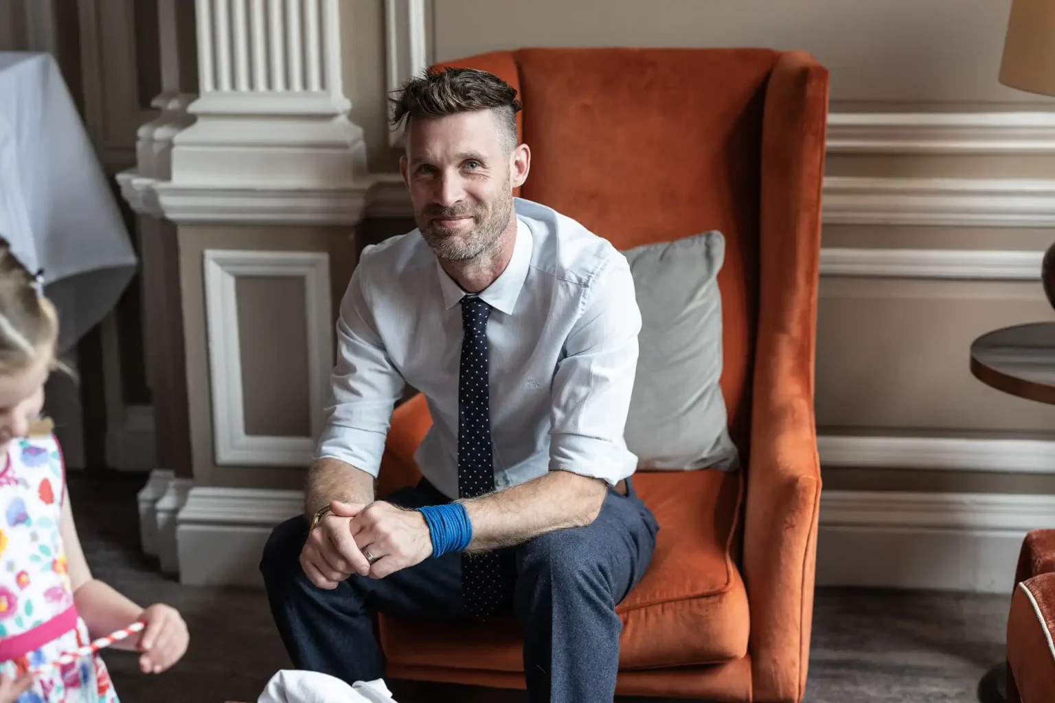 A man in a dress shirt and tie sits smiling on an orange chair, looking at a young girl who is partially visible in the foreground.