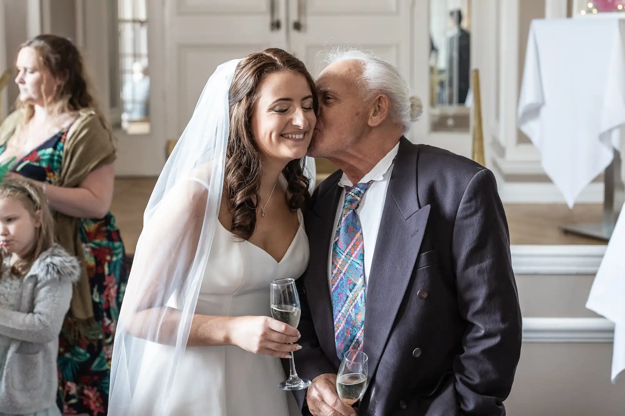 A bride in a white dress smiles as an elderly man kisses her cheek at a wedding reception, both holding champagne glasses.