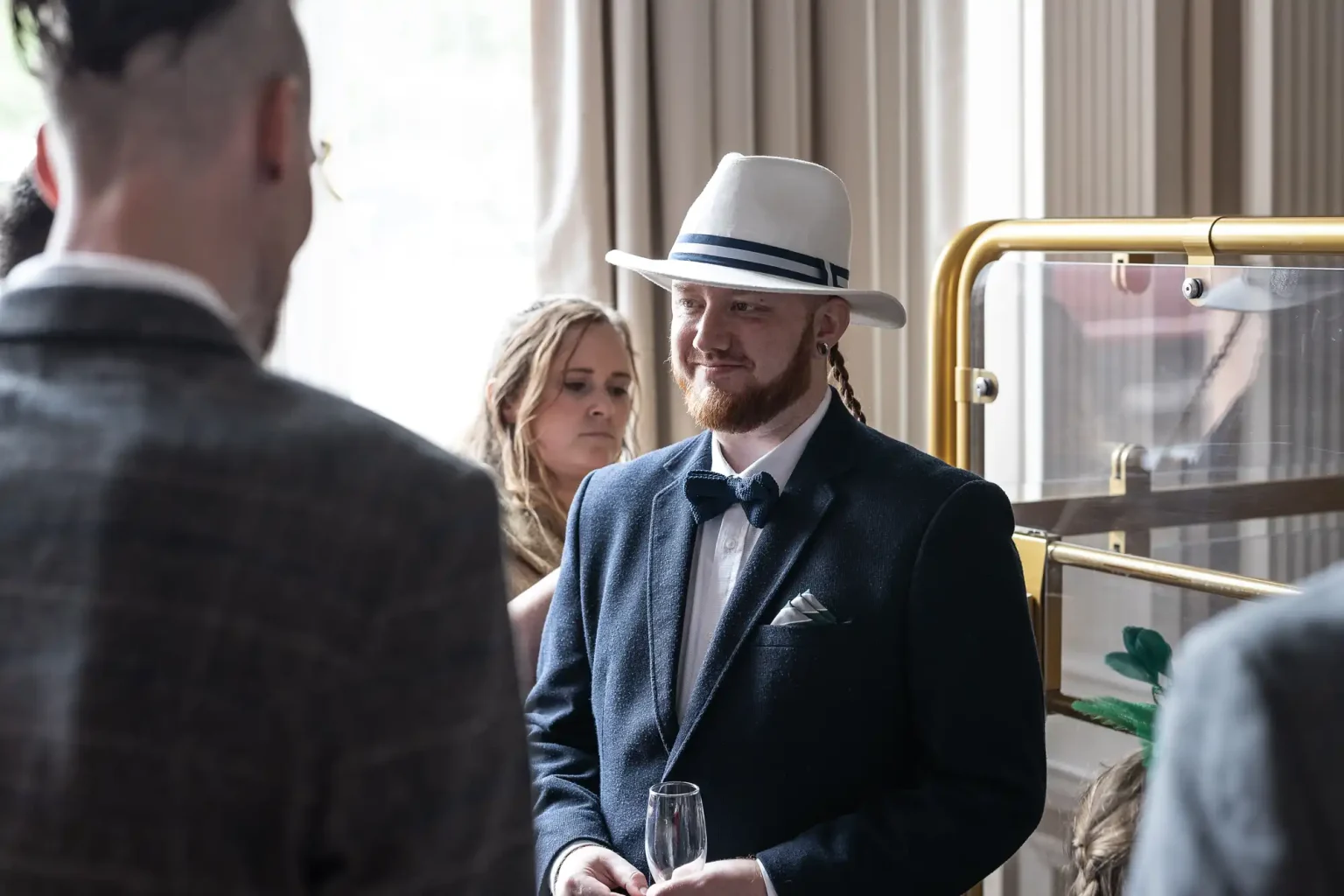 A man in a dark suit and white fedora hat holds a glass of champagne, smiling at another man at a social event.