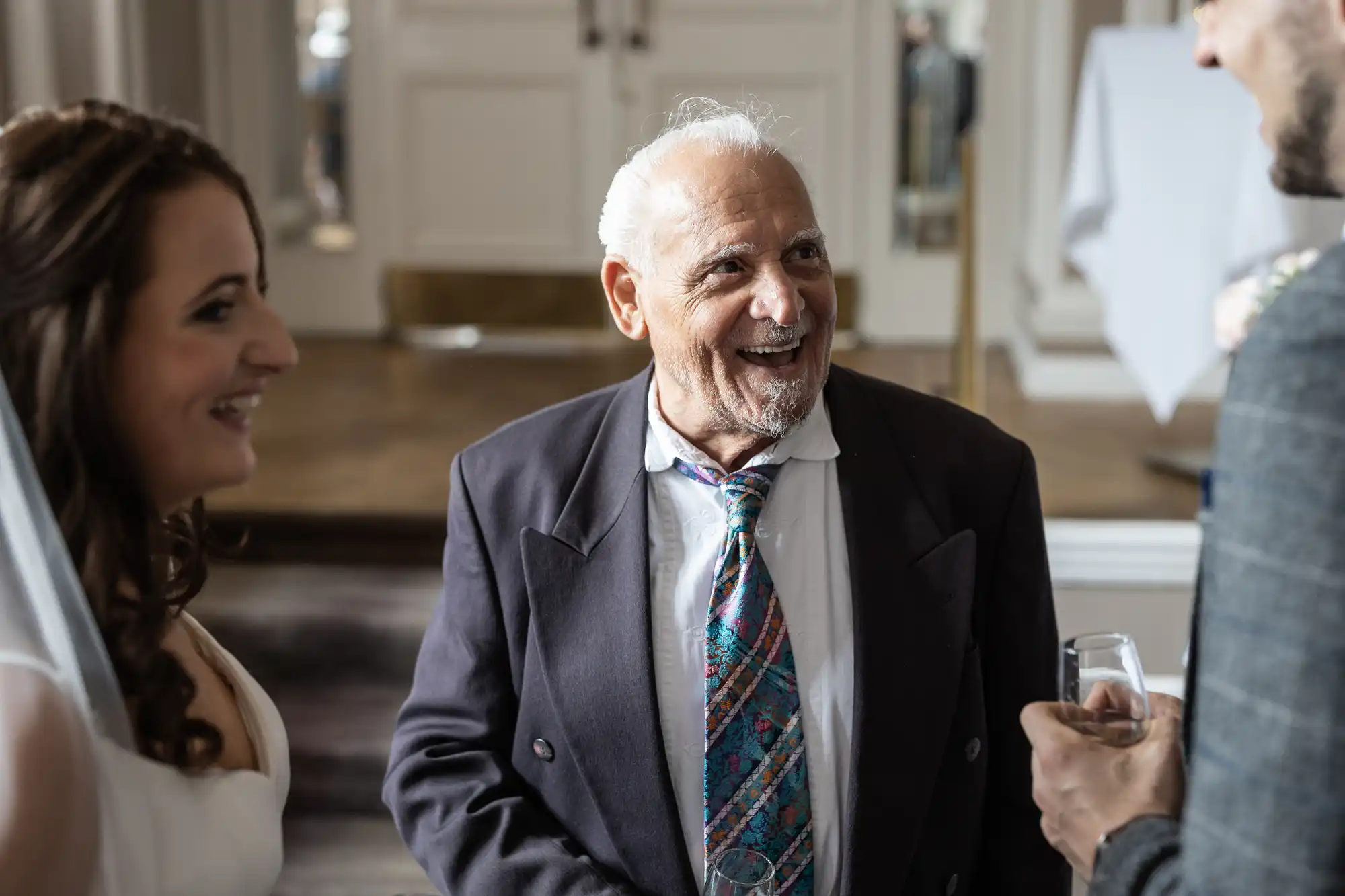 An elderly man in a suit smiling joyfully during a conversation with a bride and groom at a wedding reception.