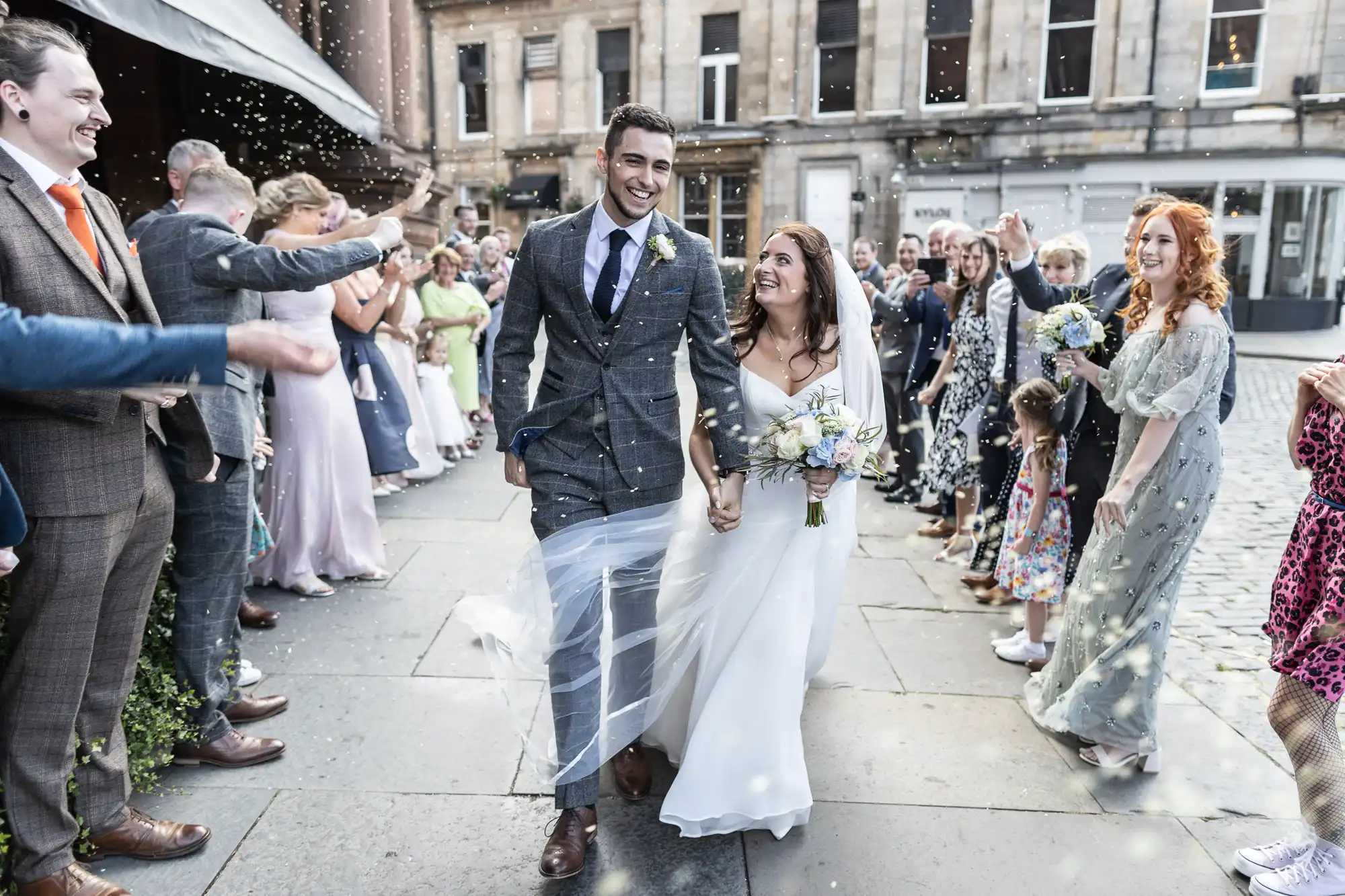 A newly married couple walks joyfully through a crowd throwing confetti on a city street.