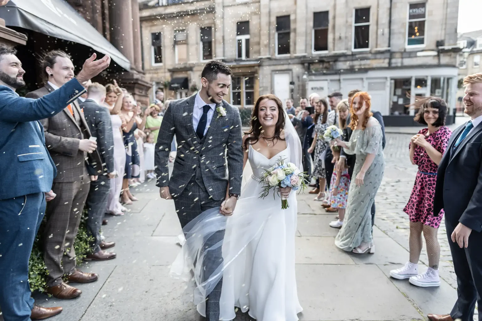 A newly married couple walks joyfully through a crowd throwing confetti on a city street.
