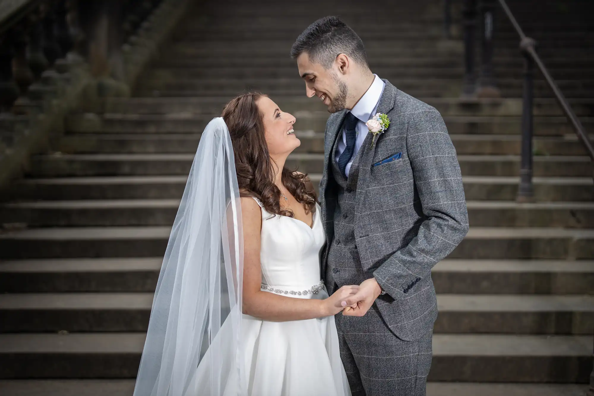 A bride in a white dress and a groom in a gray suit holding hands and gazing at each other on stone stairs.