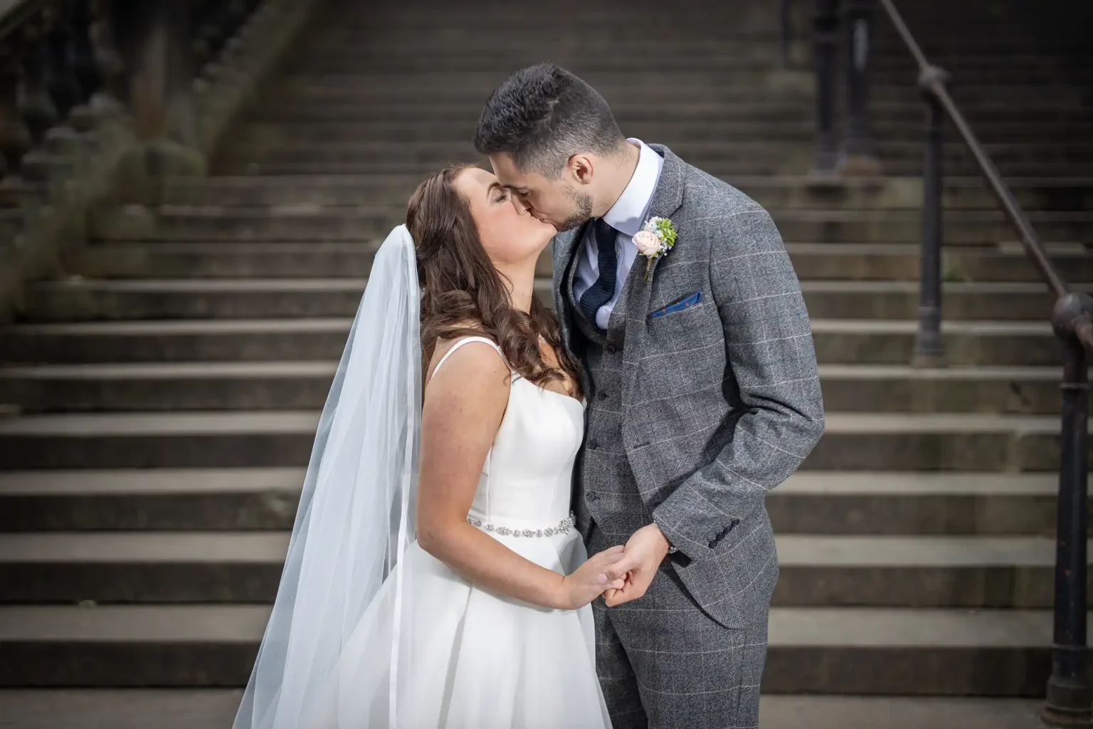 A bride and groom kissing on steps, the bride in a white dress and veil, and the groom in a gray checked suit.