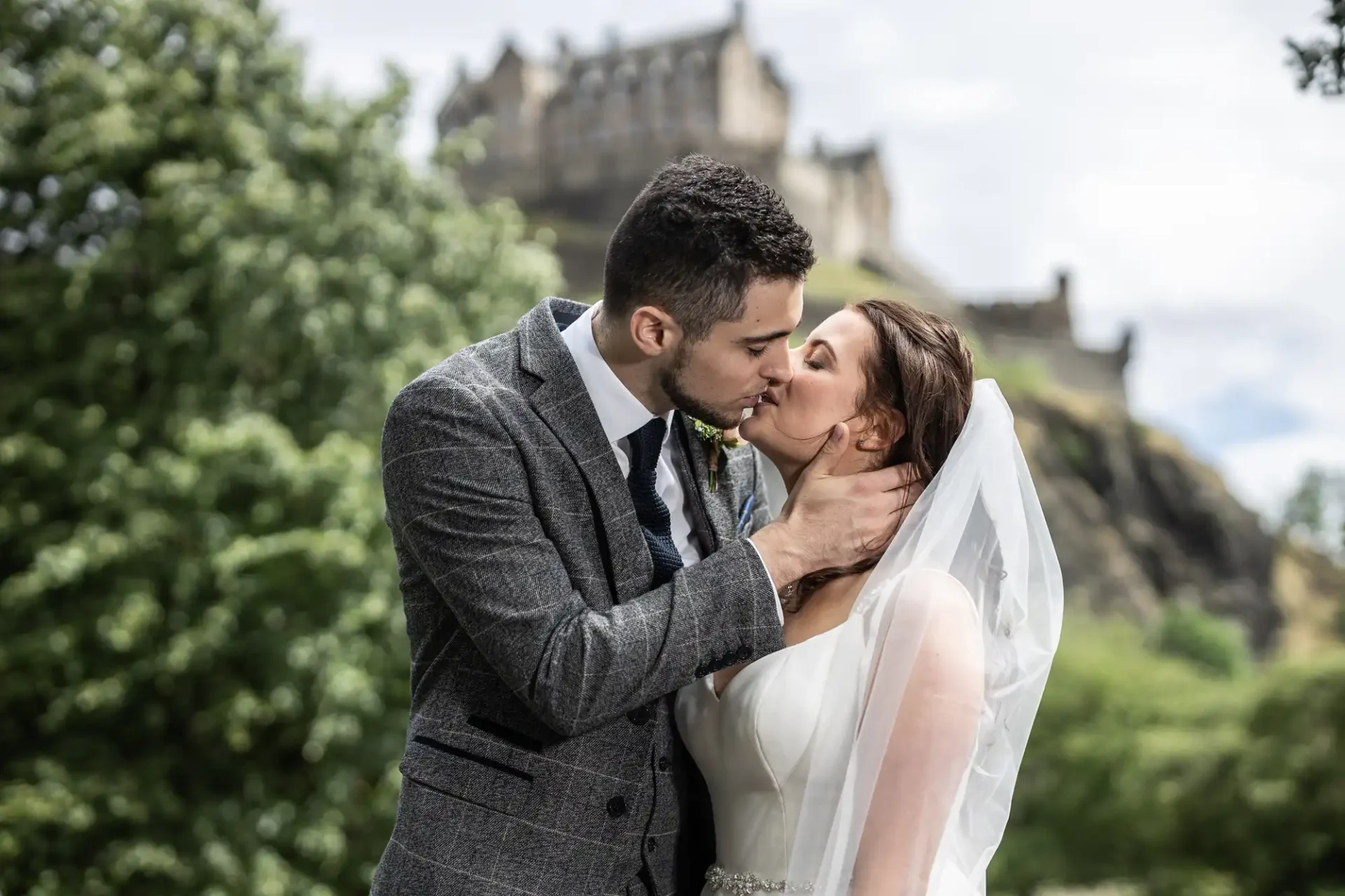A bride and groom kissing outdoors with a castle on a hill in the background.